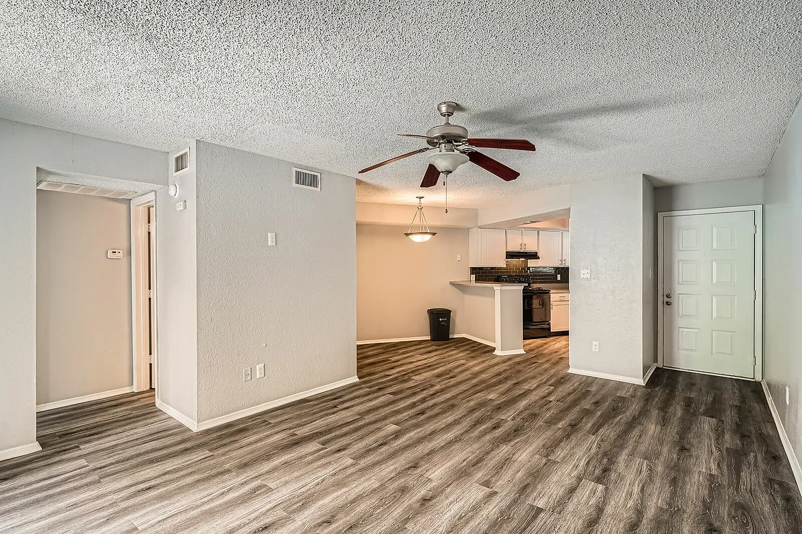 Living room with wood-style plank flooring, ceiling fan, kitchen entrance, white walls