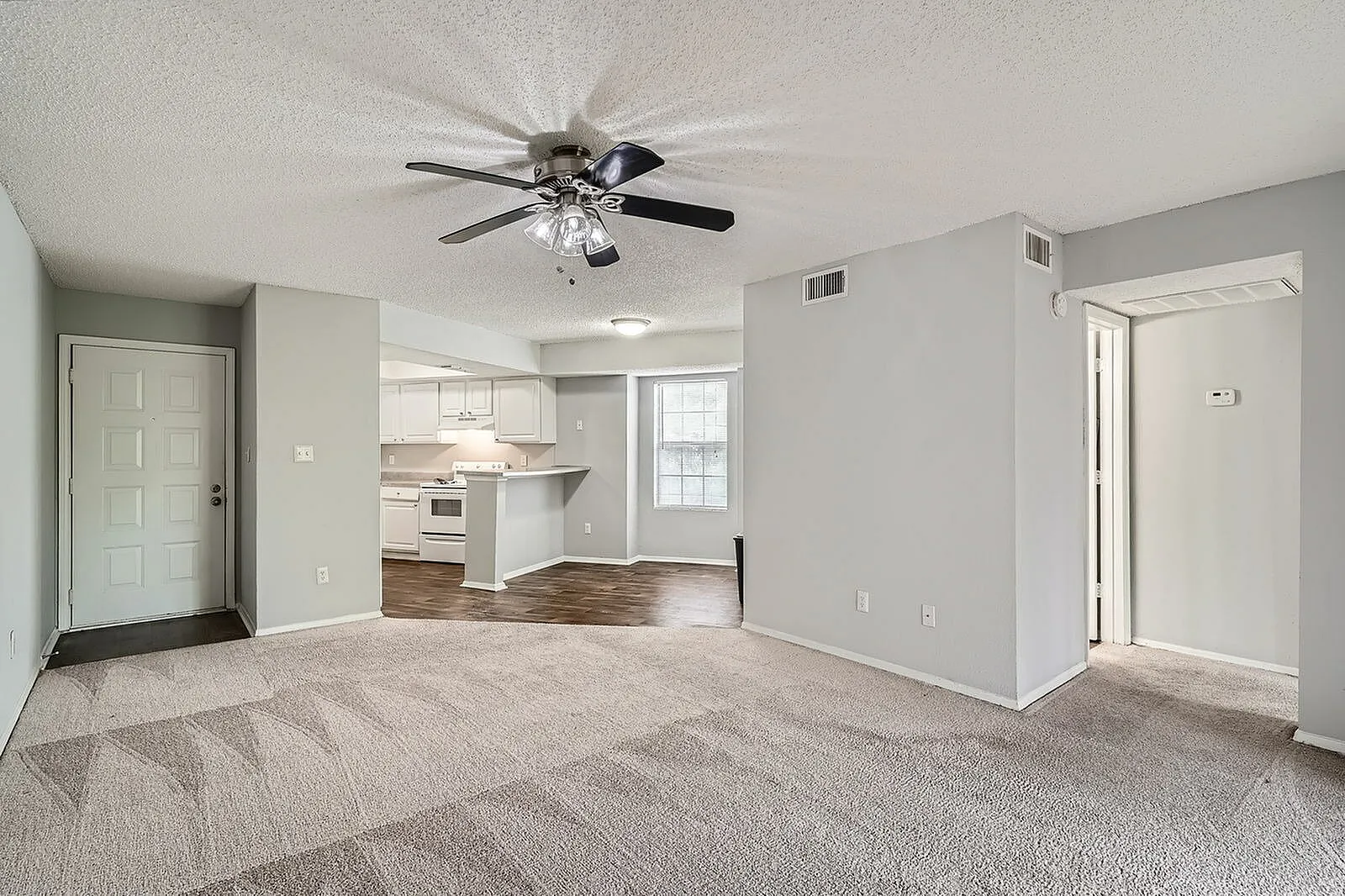 Living room, wood-style plank flooring, ceiling fan, kitchen with white cabinetry visible