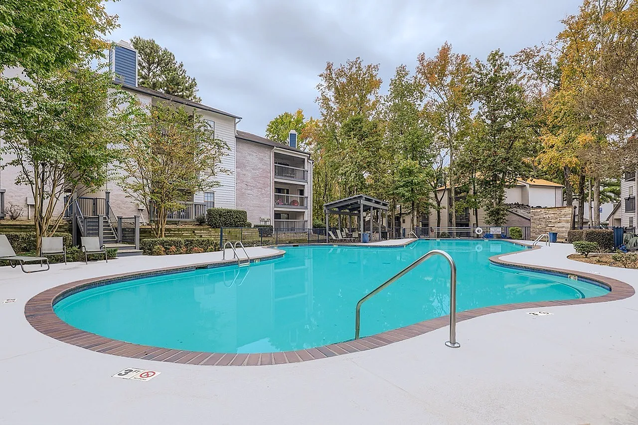 Kidney-shaped pool with turquoise water, metal handrail, brick decking, apartment buildings with trees
