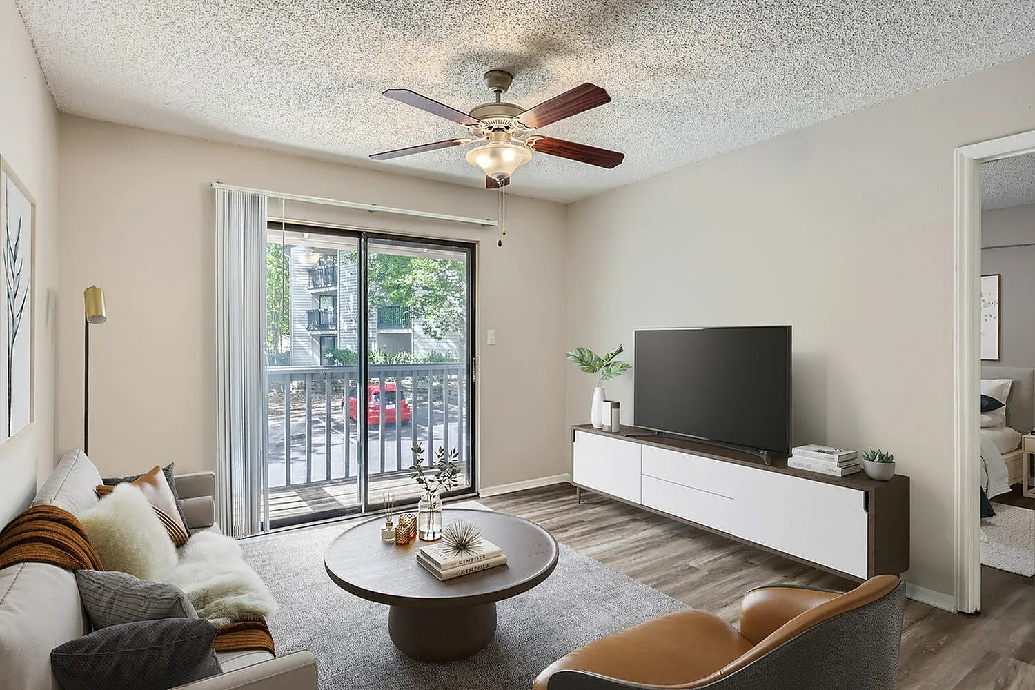 Living room with wood-style plank flooring, ceiling fan, sliding glass balcony door, gray sofa, leather chair, flat-screen TV mounted on media console