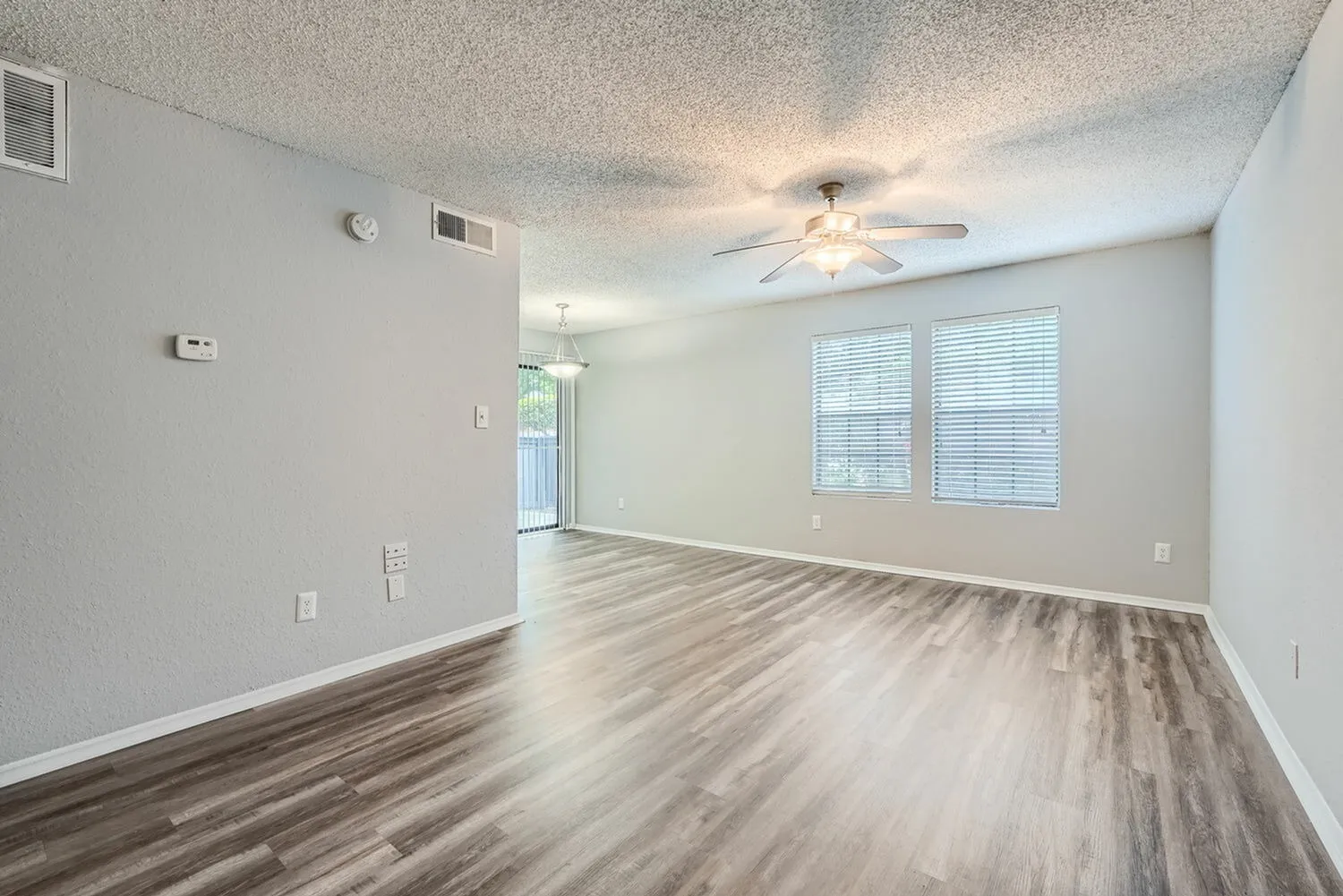 Living room, wood-style plank flooring, ceiling fan, large windows, entry doorway