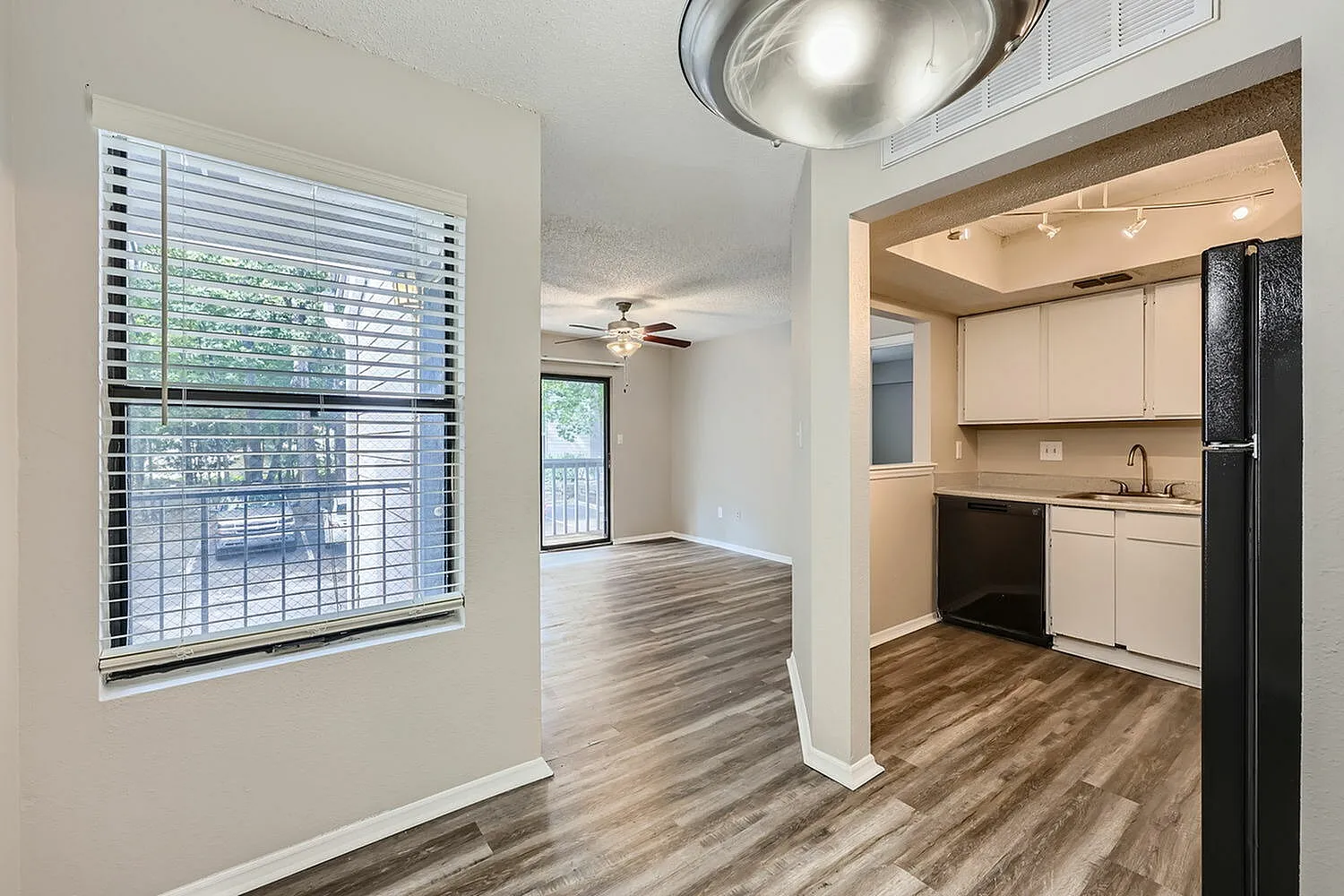 Living room with wood-style plank flooring, large window with blinds, kitchen with white cabinetry and black appliances