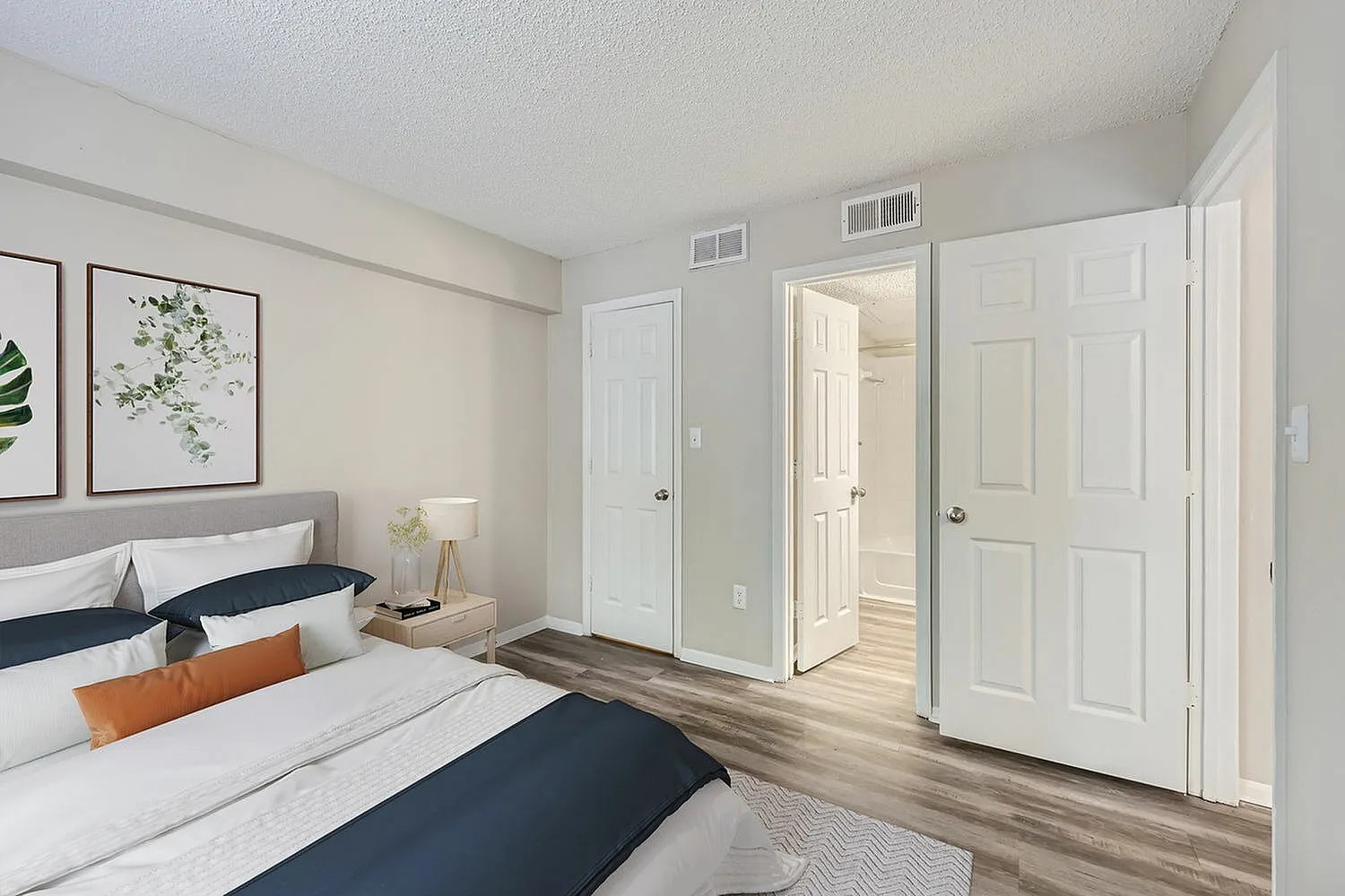 Primary bedroom, wood-style plank flooring, white bedding, orange accents, ensuite bathroom visible