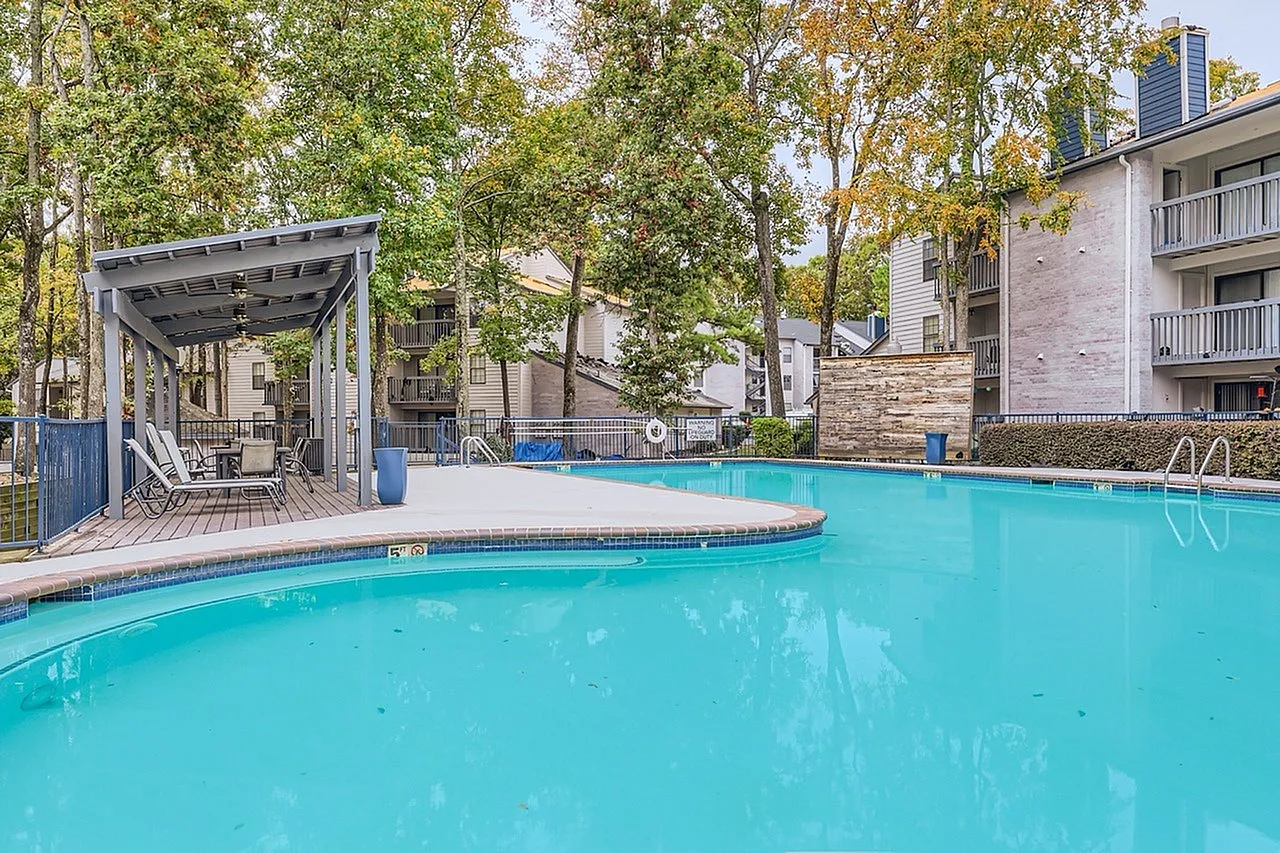 Resort-style swimming pool, lounge seating under pergola, brick building exterior, mature trees