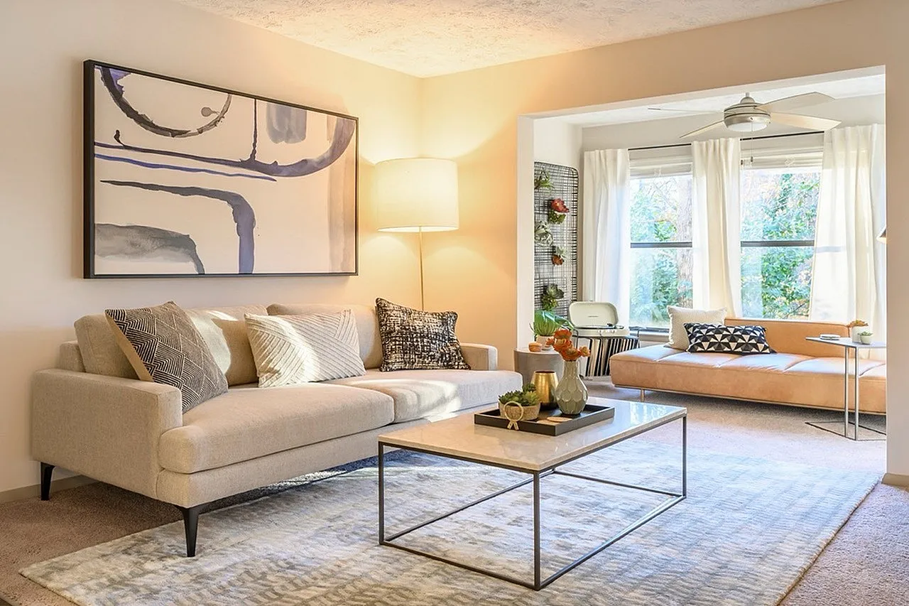 Living room, cream upholstered sofa, framed abstract art, metal coffee table, adjoining den with wood-style plank flooring