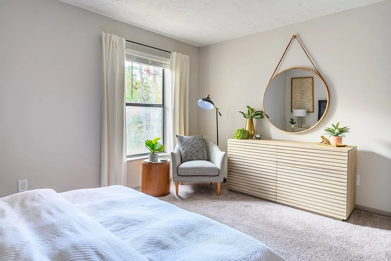 Primary bedroom, light wood dresser, round hanging mirror, casual accent chair, natural light