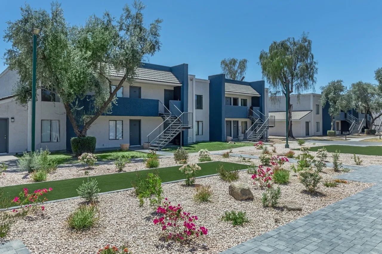 Apartment complex exterior, multi-story units, landscaped courtyard with flowering shrubs, gravel and paved walkways, clear sky