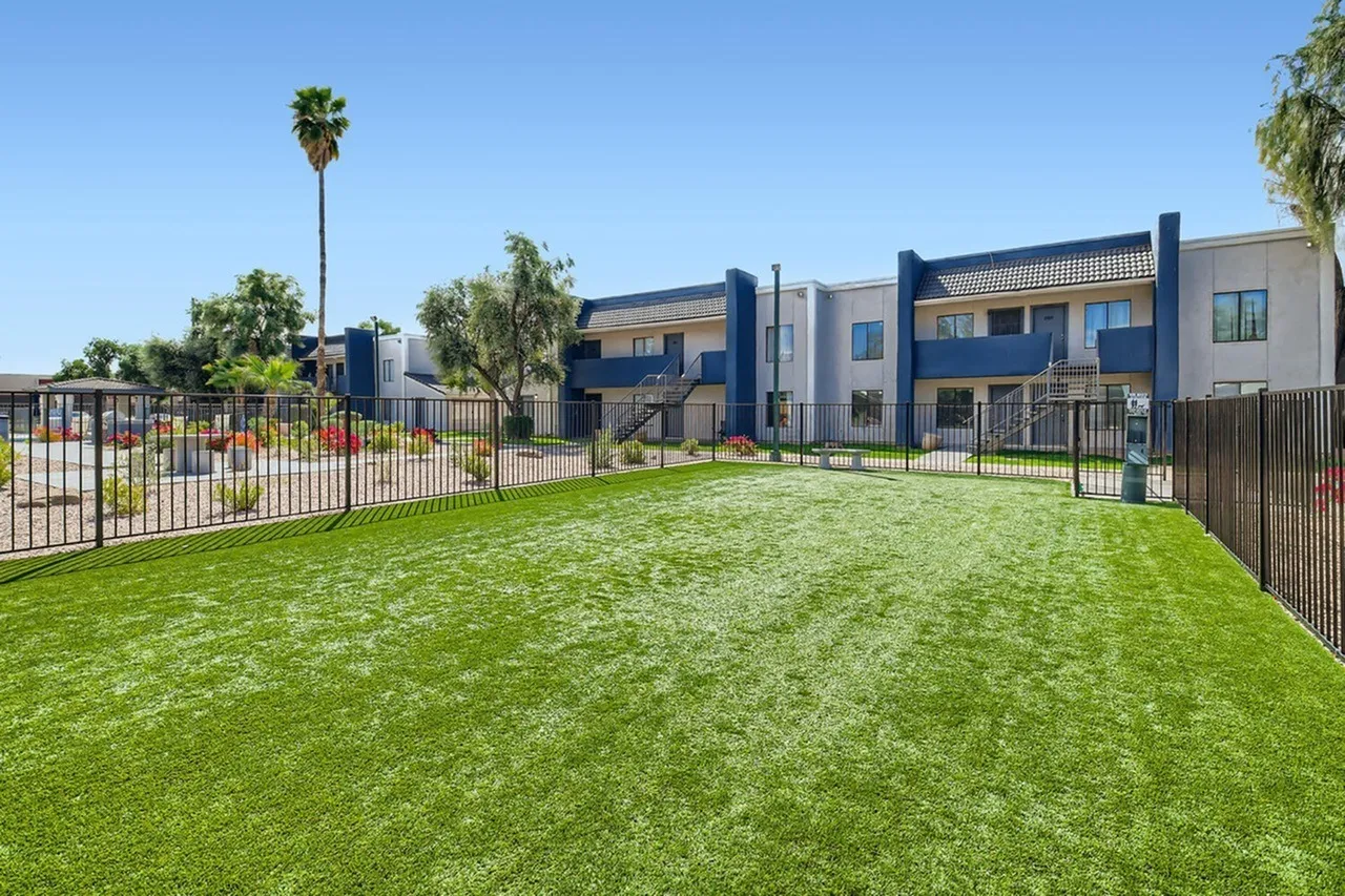 Community courtyard with grass lawn, metal fencing, modern apartment buildings, trees, blue sky