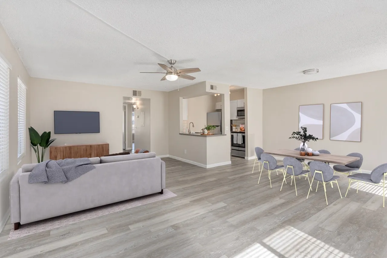 Open living space with light gray sofa, wood-style plank flooring, ceiling fan, kitchen with white cabinetry visible in background