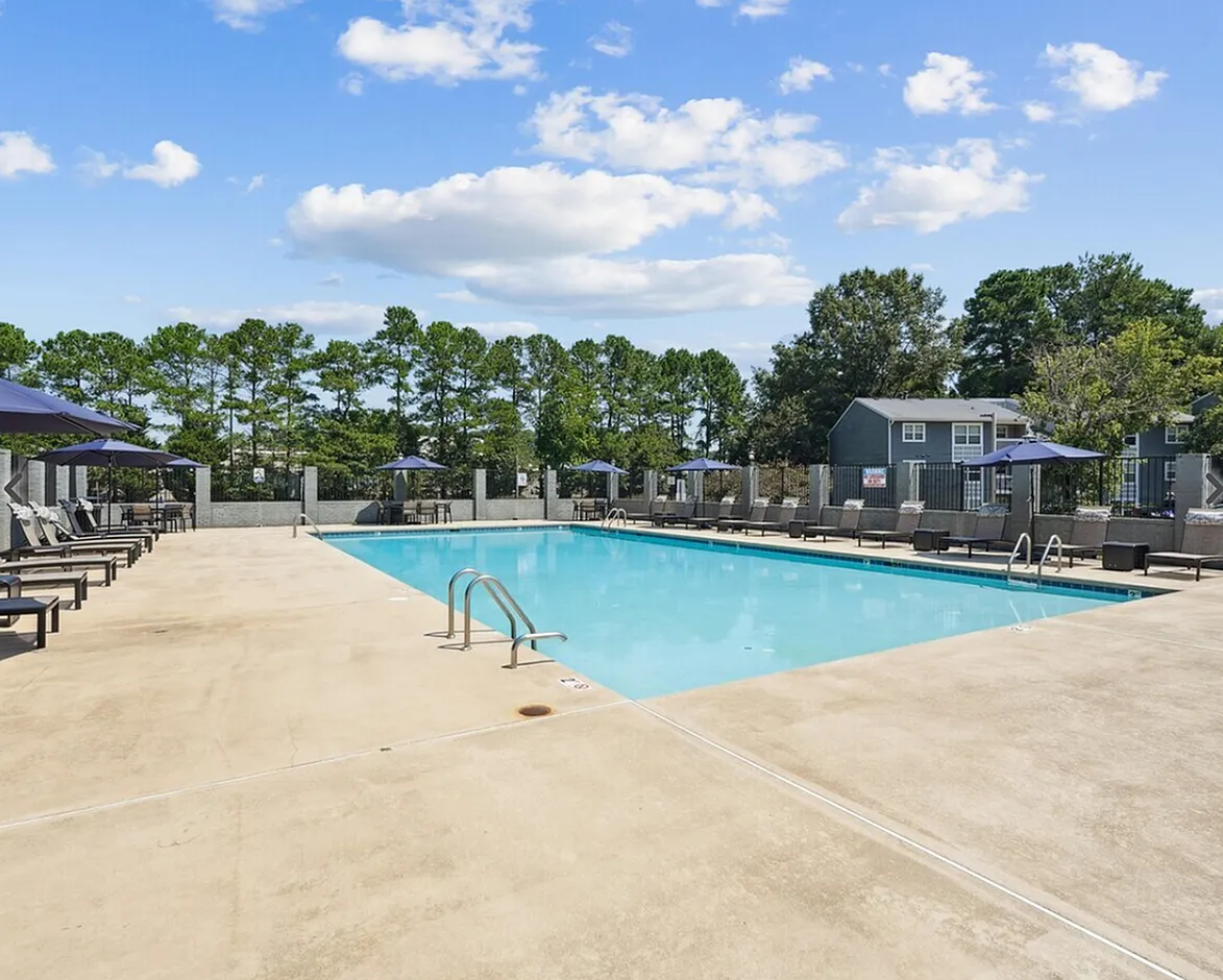 Swimming pool, lounge chairs, concrete deck, gray pool houses, tree line
