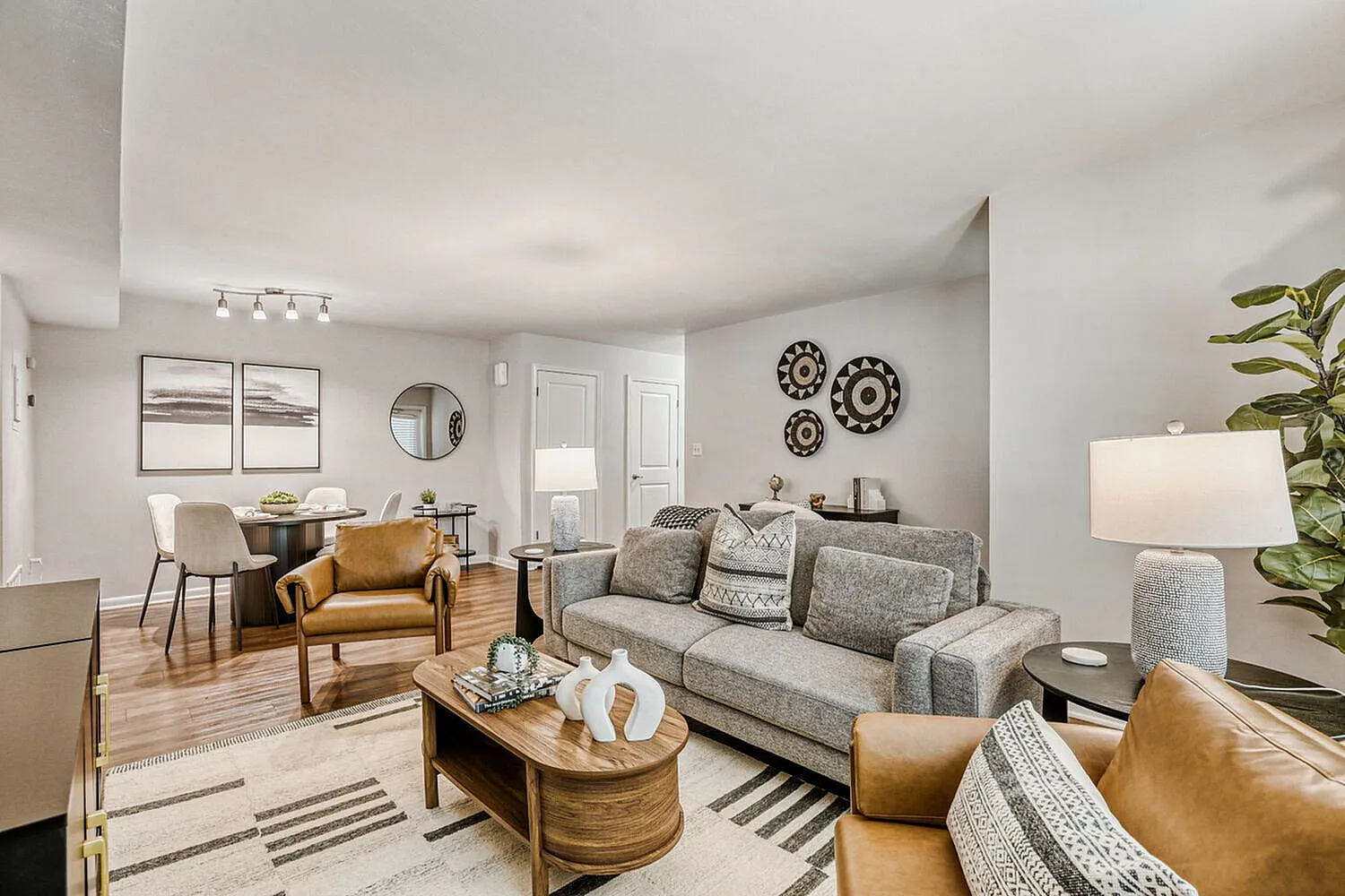 Living room with wood-style plank flooring, gray upholstered sofa, caramel leather accent chairs, wooden coffee table, wall-mounted gears, pendant light