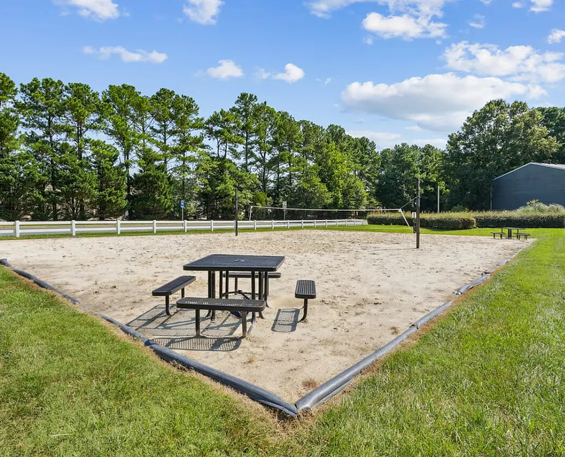 Sand volleyball court, picnic table with benches, white fence, trees, grass lawn