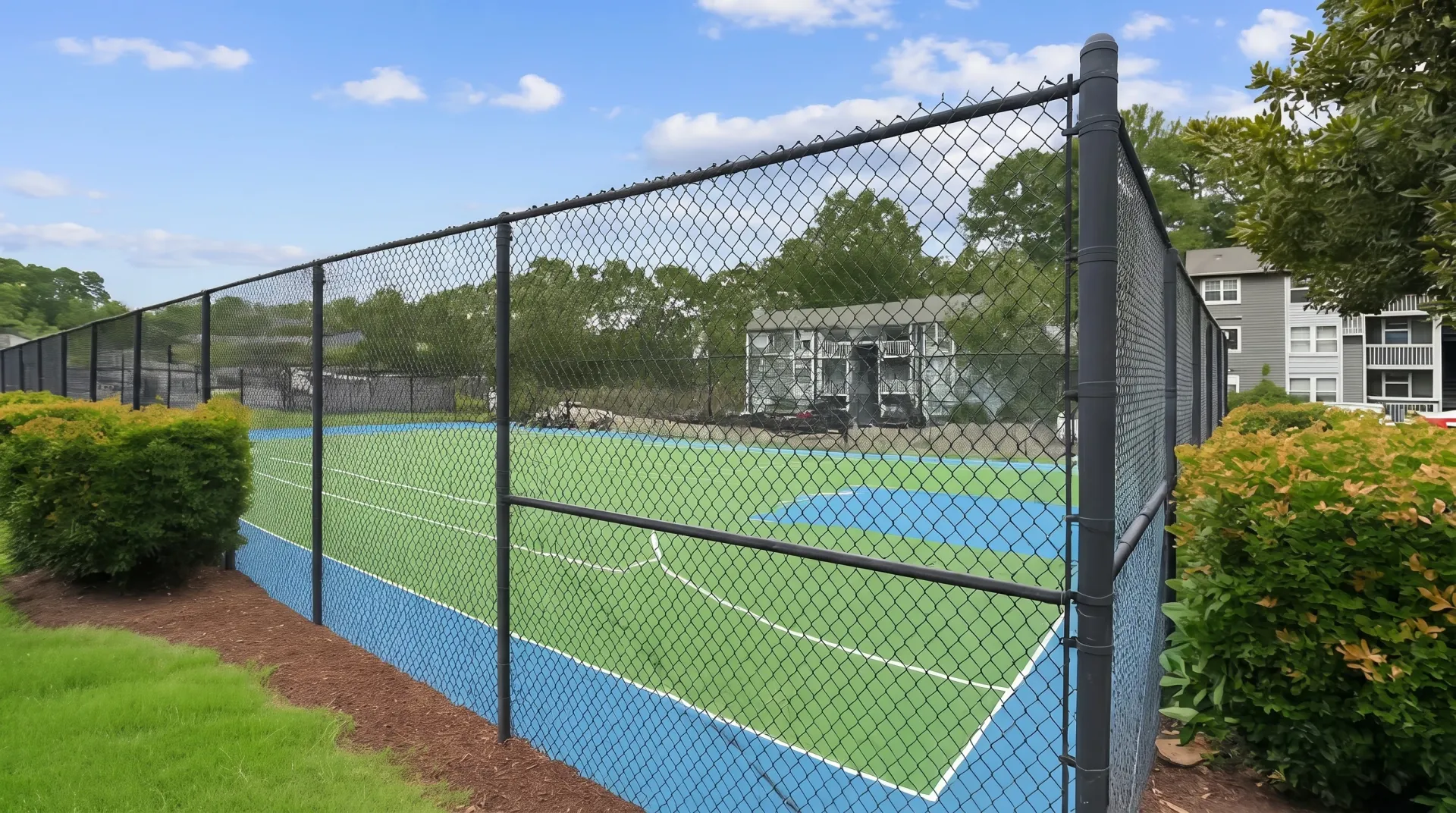 Tennis court, black chain-link fencing, blue hard court surface, hedge landscaping, residential building