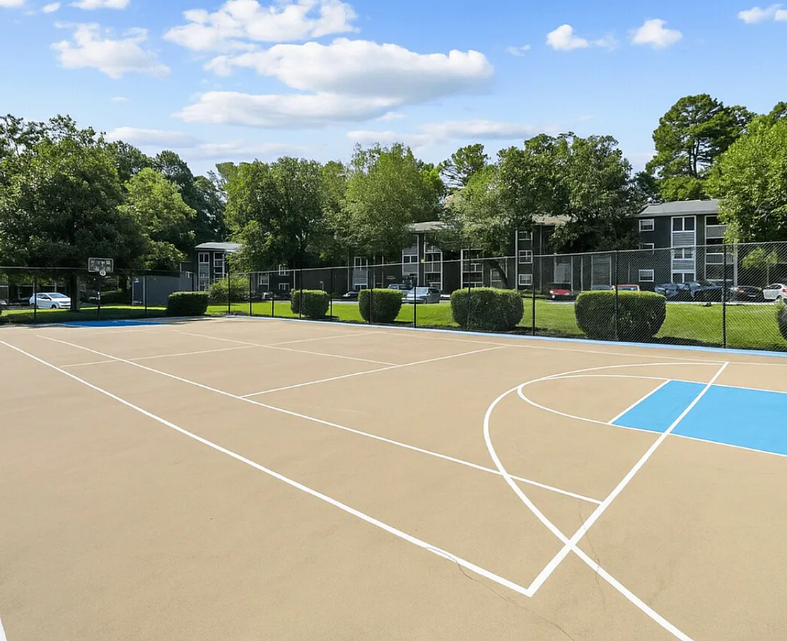 Multi-sport court with white line markings, blue basketball key, tan playing surface, residential buildings and landscaping in background