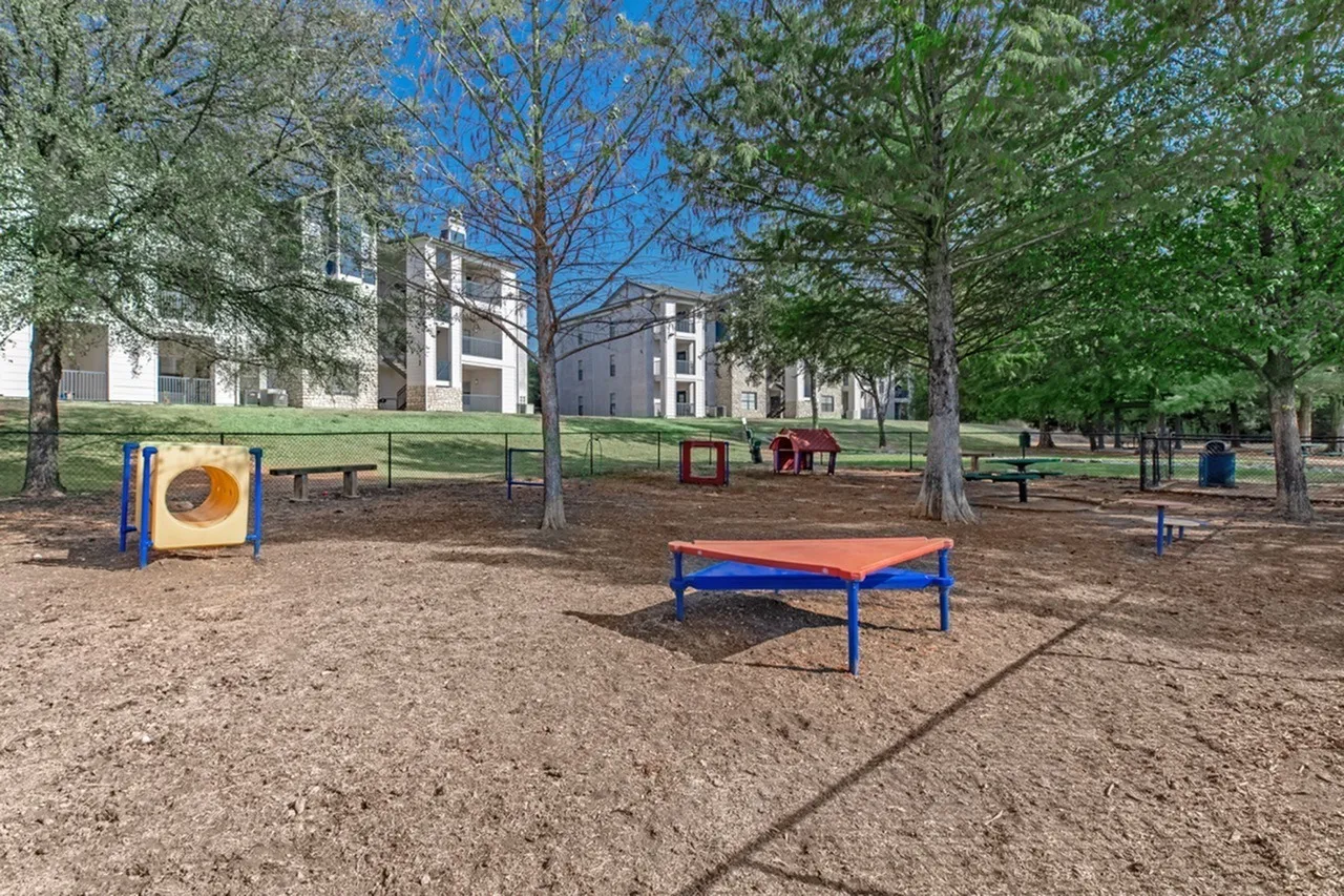 Playground with orange trampoline, yellow tunnel climber, blue picnic table, mature trees, brick buildings in background