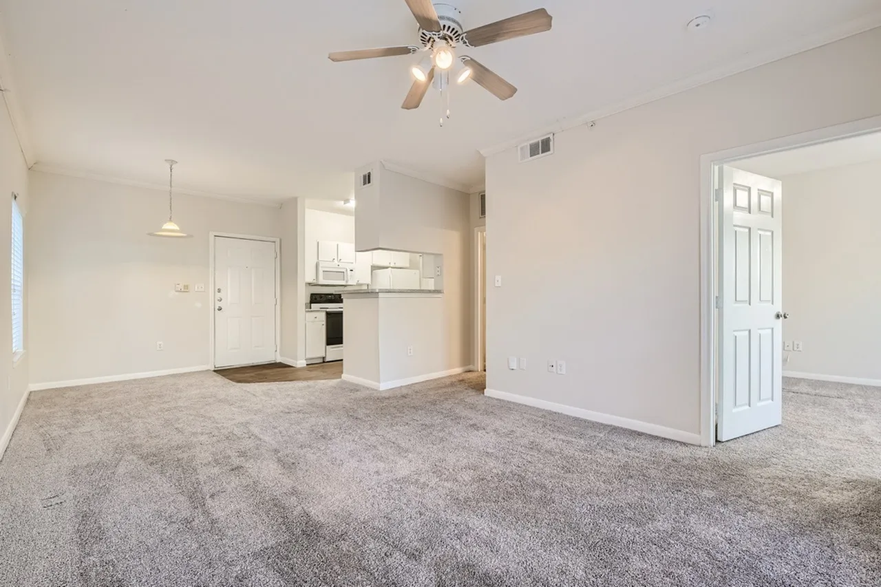 Living room with gray carpet, ceiling fan, white kitchen with white cabinetry and stainless appliances, entry door
