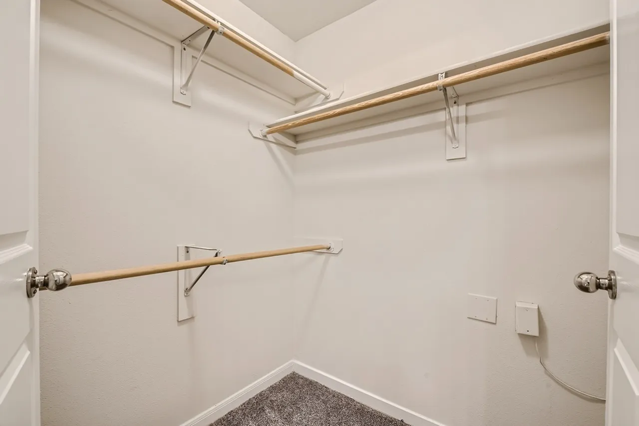 Walk-in closet, wooden hanging rods, white walls, gray carpet, wall outlets