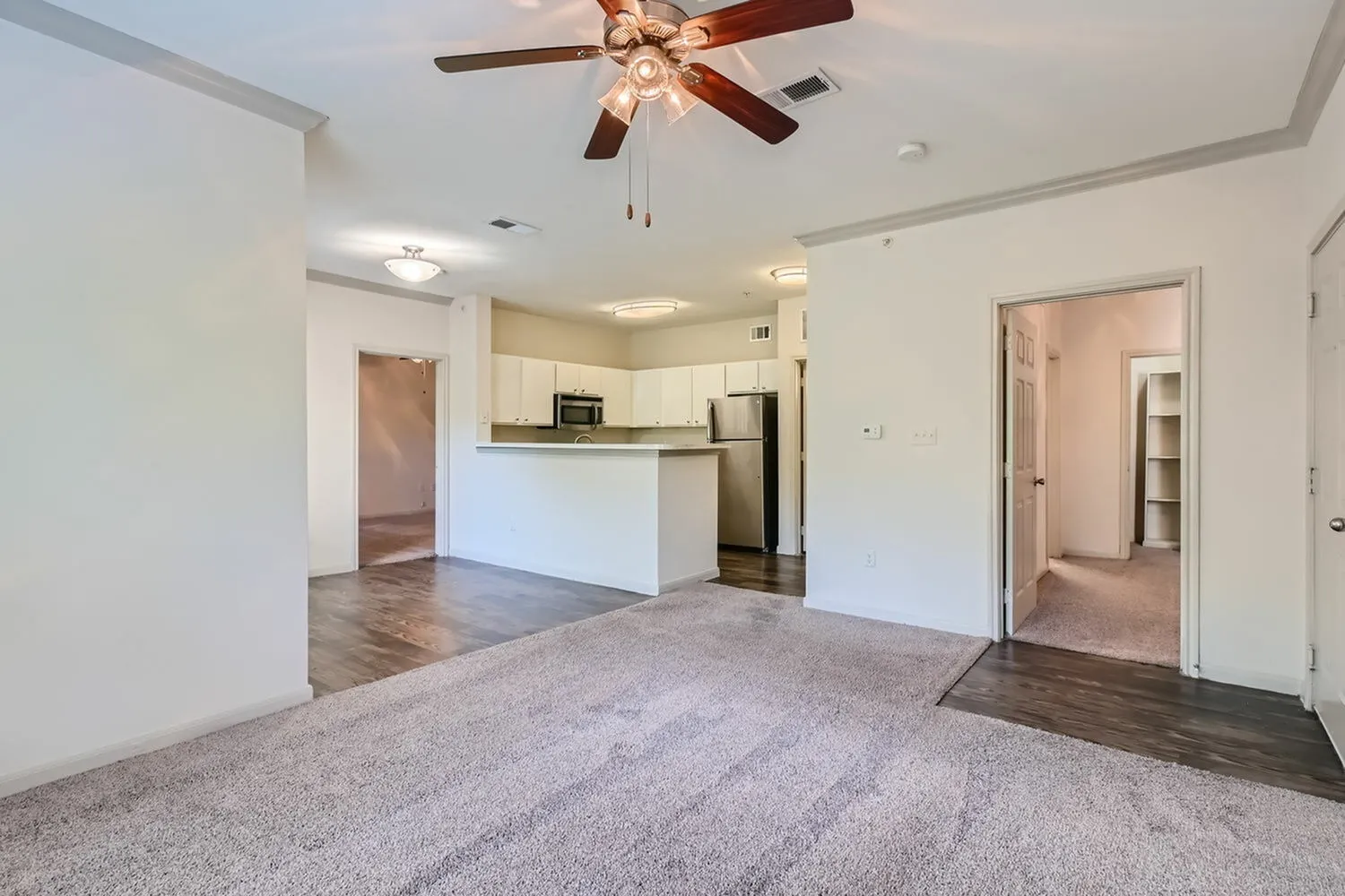 Open living room with wood-style plank flooring, ceiling fan, kitchen with white cabinetry and stainless appliances, white island, gray carpet in foreground