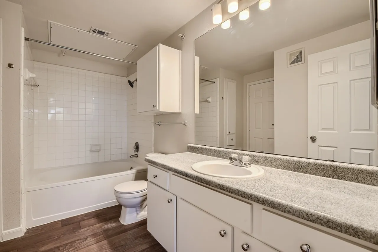 Primary bathroom, gray quartz countertops, white vanity cabinetry, white subway tile shower surround, wood-style plank flooring