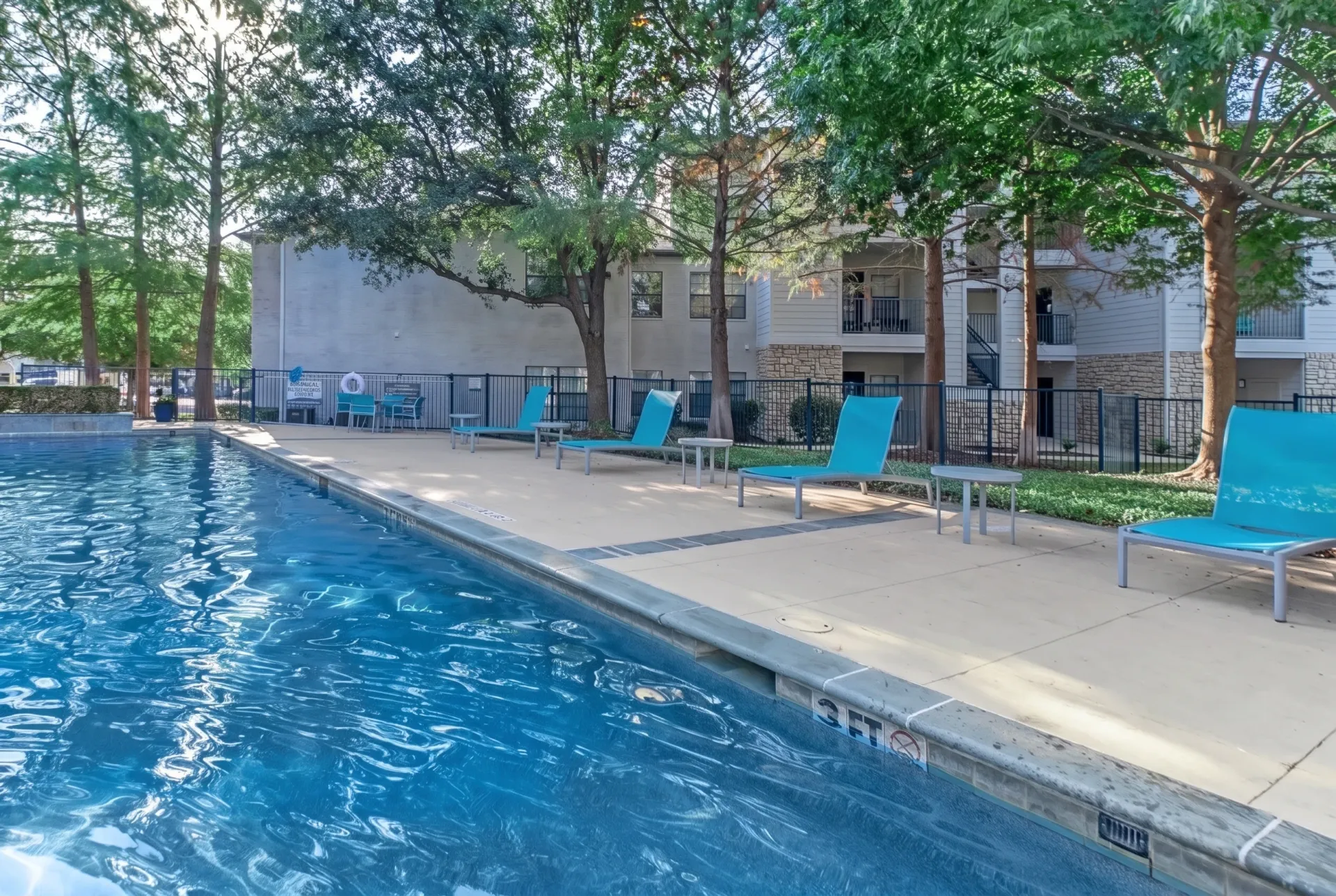 Swimming pool, blue lounge chairs, concrete patio, apartment building, mature trees
