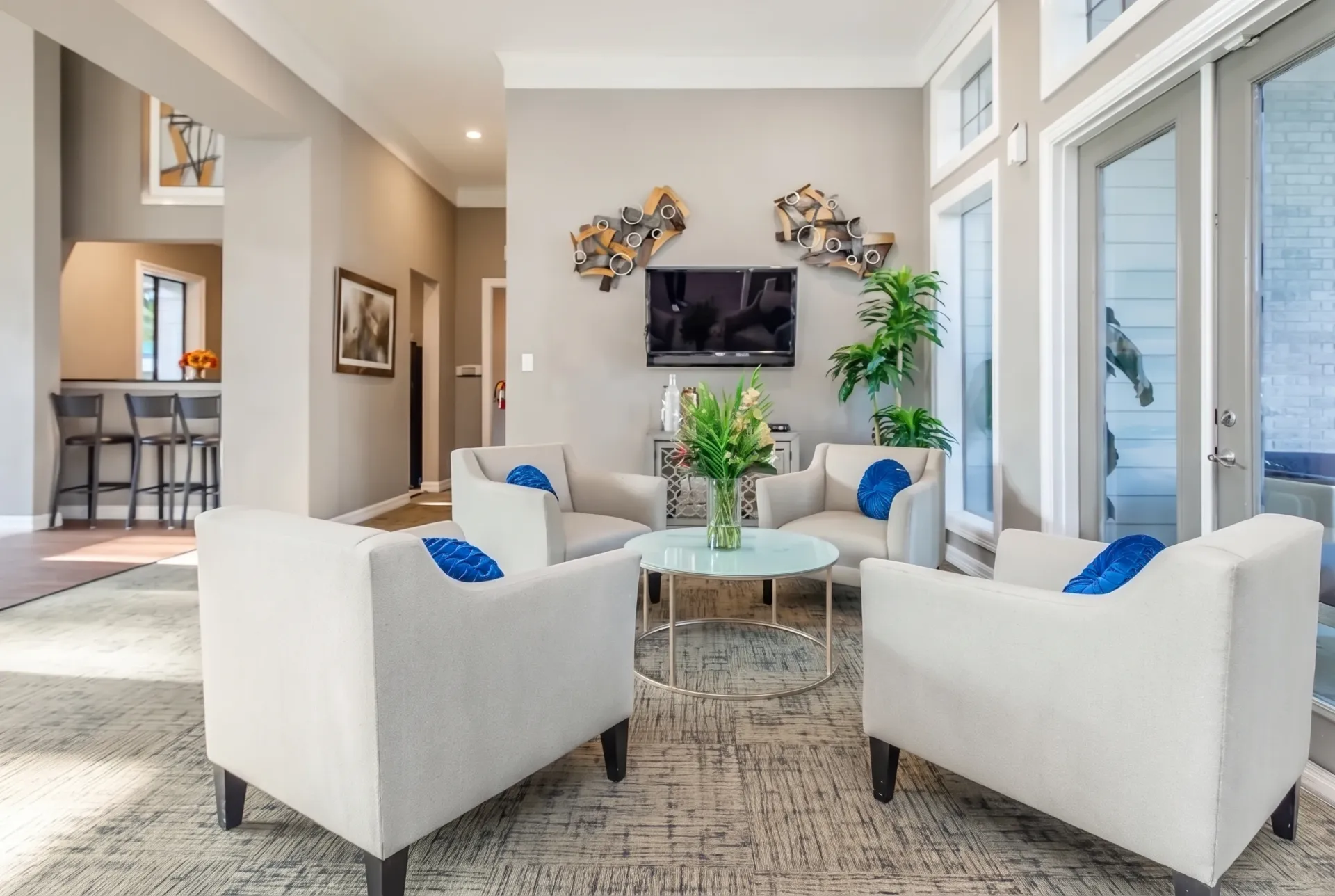 Lounge area, cream upholstered chairs with blue accent pillows, wood-style plank flooring, mounted TV, glass doors