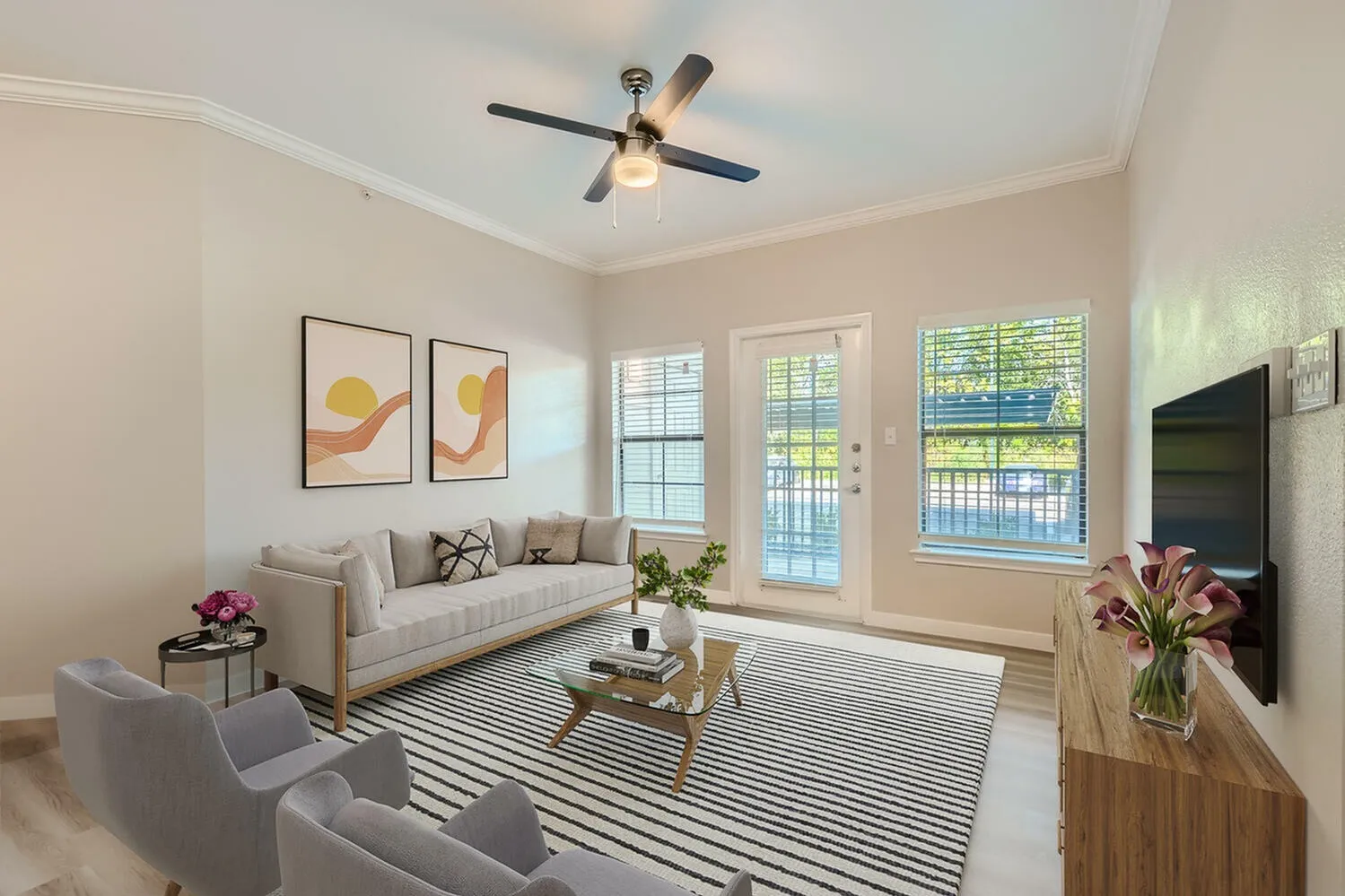 Living room with wood-style plank flooring, striped area rug, ceiling fan, neutral sectional sofa, glass doors with shutters, framed artwork, television, side table with flowers