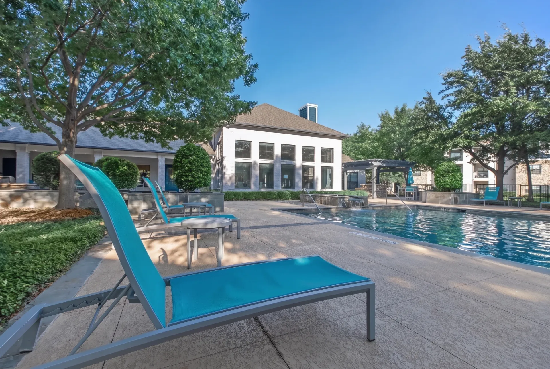 Pool area with blue lounge chairs, resort-style seating, mature trees, clubhouse