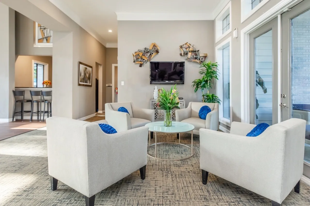 Lounge area, cream upholstered chairs with blue accent pillows, wood-style plank flooring, mounted TV, glass doors