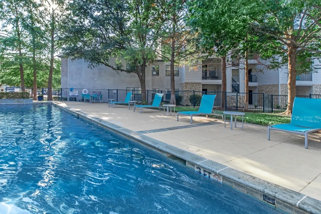 Swimming pool, blue lounge chairs, concrete patio, apartment building, mature trees