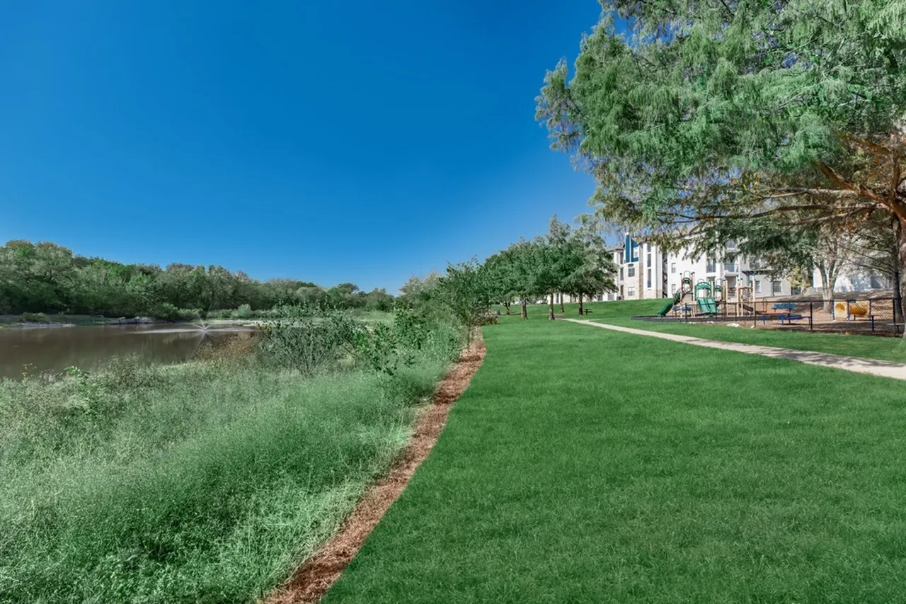 Waterfront park area, manicured lawn, mature trees, building exterior, playground