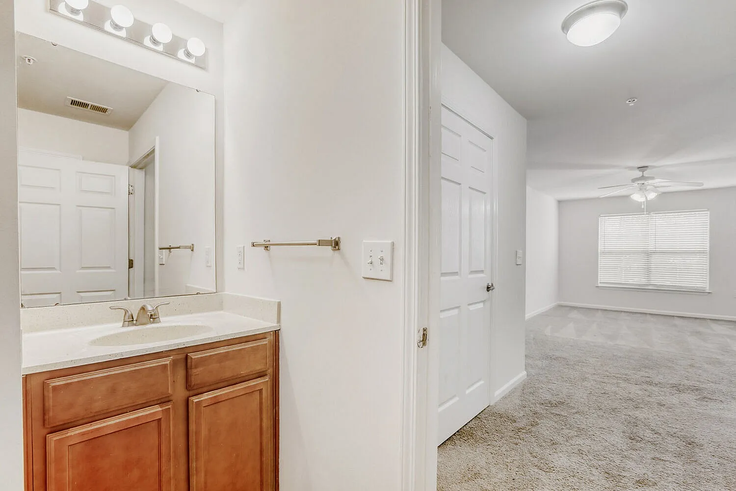 Primary bathroom with wood cabinetry vanity, white countertop, large mirror, ceiling lighting, bedroom with ceiling fan and wood-style plank flooring visible beyond