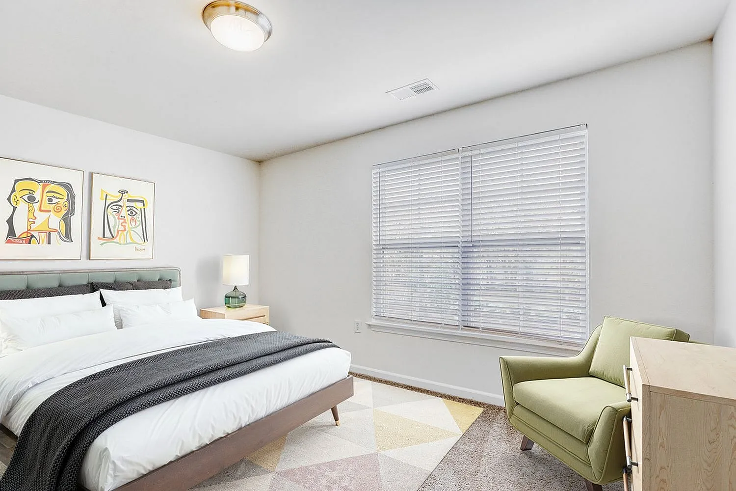 Primary bedroom, bed with white and gray bedding, abstract wall art, large windows with blinds, green upholstered chair