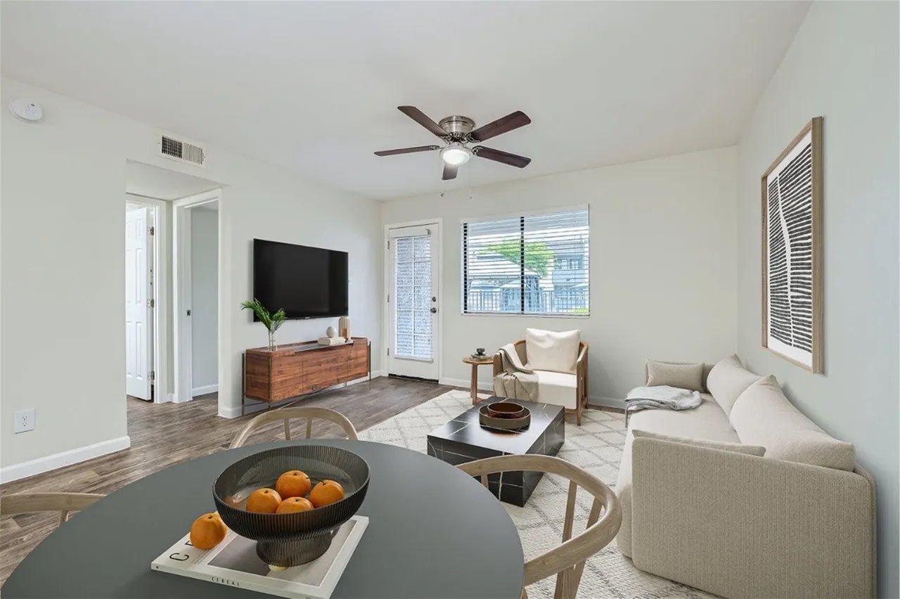 Living room with wood-style plank flooring, ceiling fan, TV mounted on dresser, windows with blinds, upholstered seating, round dining table