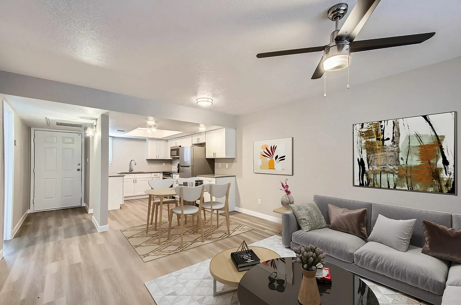 Open concept living and kitchen area, wood-style plank flooring, gray upholstered sofa, ceiling fan with light fixture, white cabinetry, stainless appliances