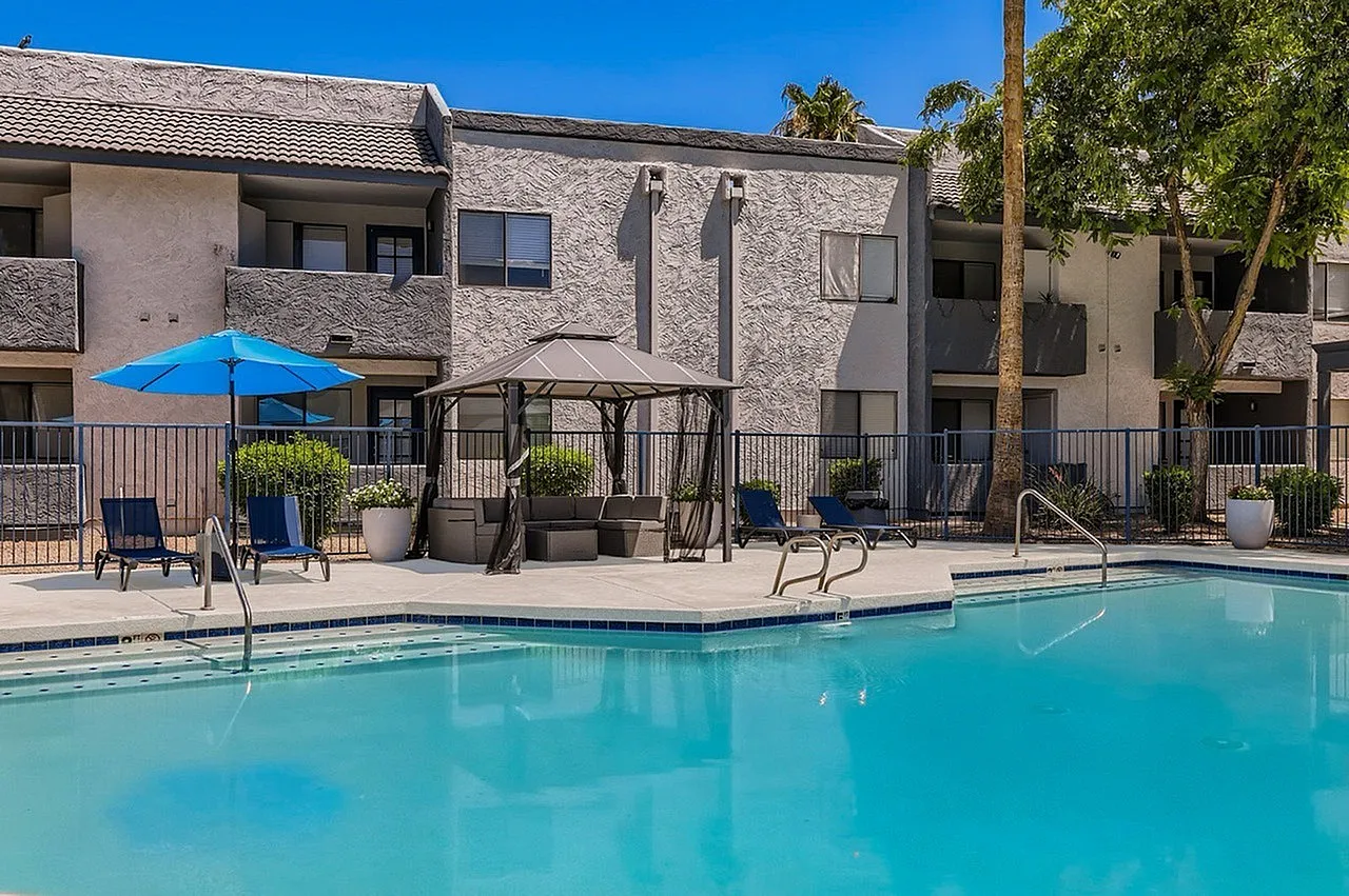 Pool area, crystal clear turquoise water, blue umbrella, lounge chairs, fenced patio, desert landscaping