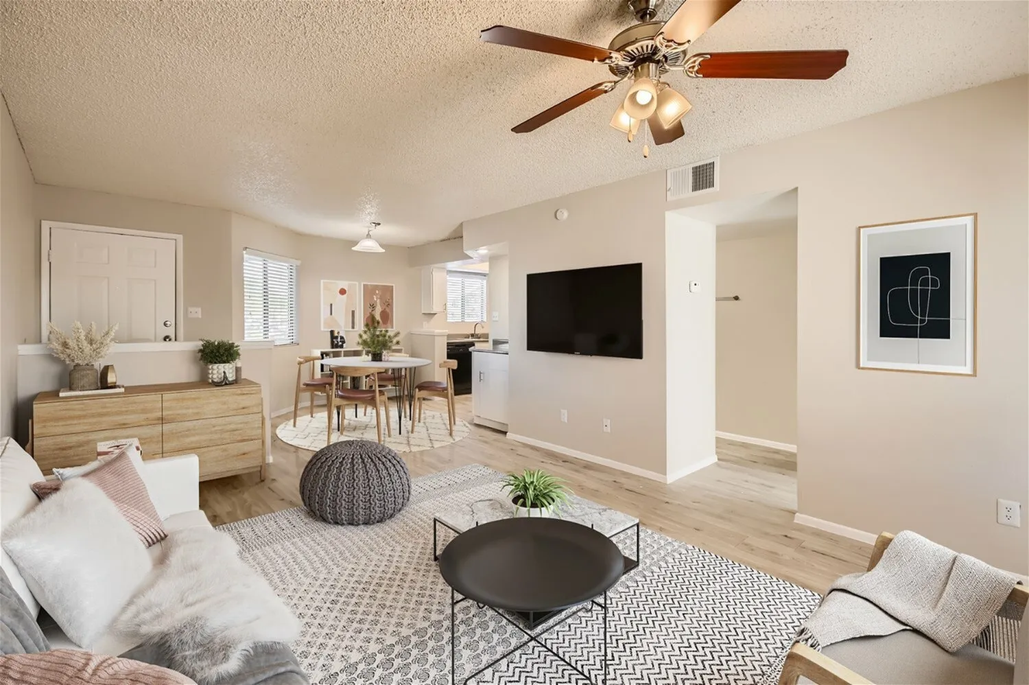 Living room with wood-style plank flooring, ceiling fan, TV wall, dining table, wood dresser, woven pouf ottoman