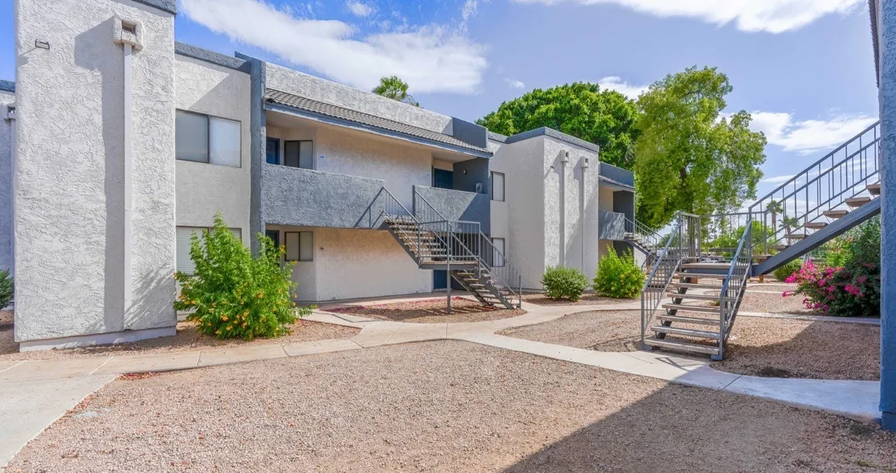 Modern multi-story residential building, concrete and stucco exterior, metal railings, gravel courtyard, landscaping