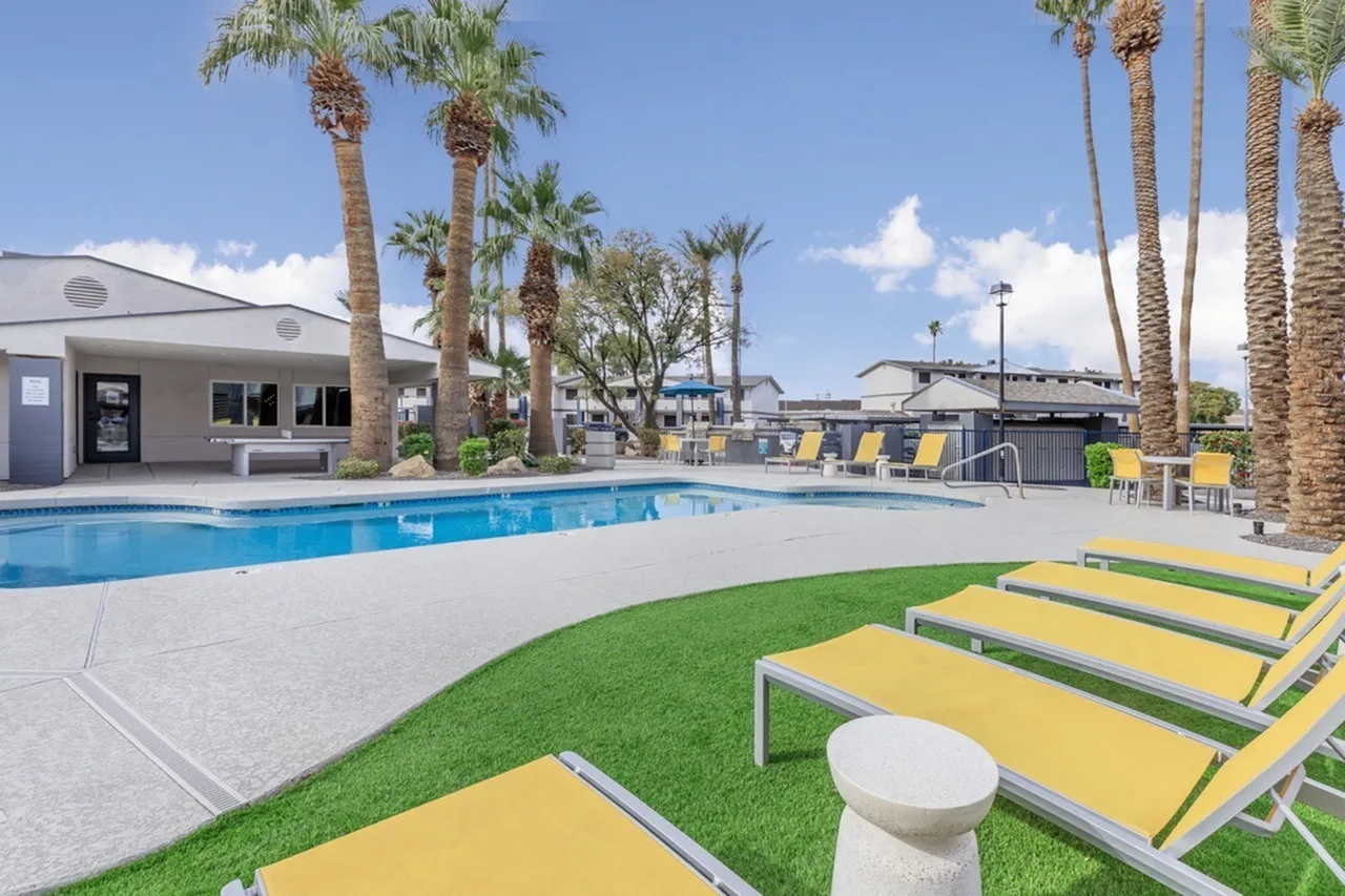 Pool deck with yellow lounge chairs, green lawn, tall palm trees, pool house building, clear blue sky