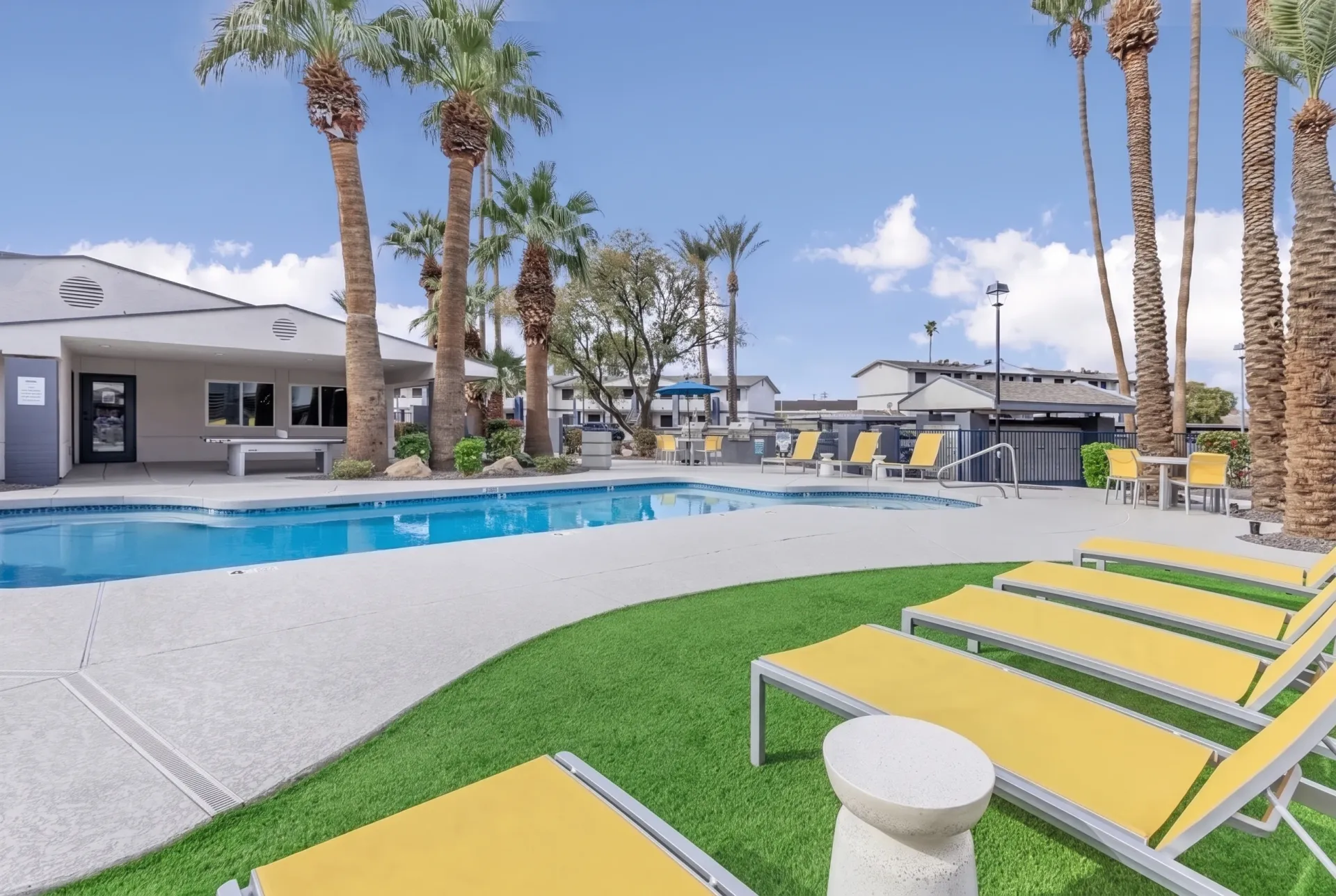 Pool deck with yellow lounge chairs, green lawn, tall palm trees, pool house building, clear blue sky