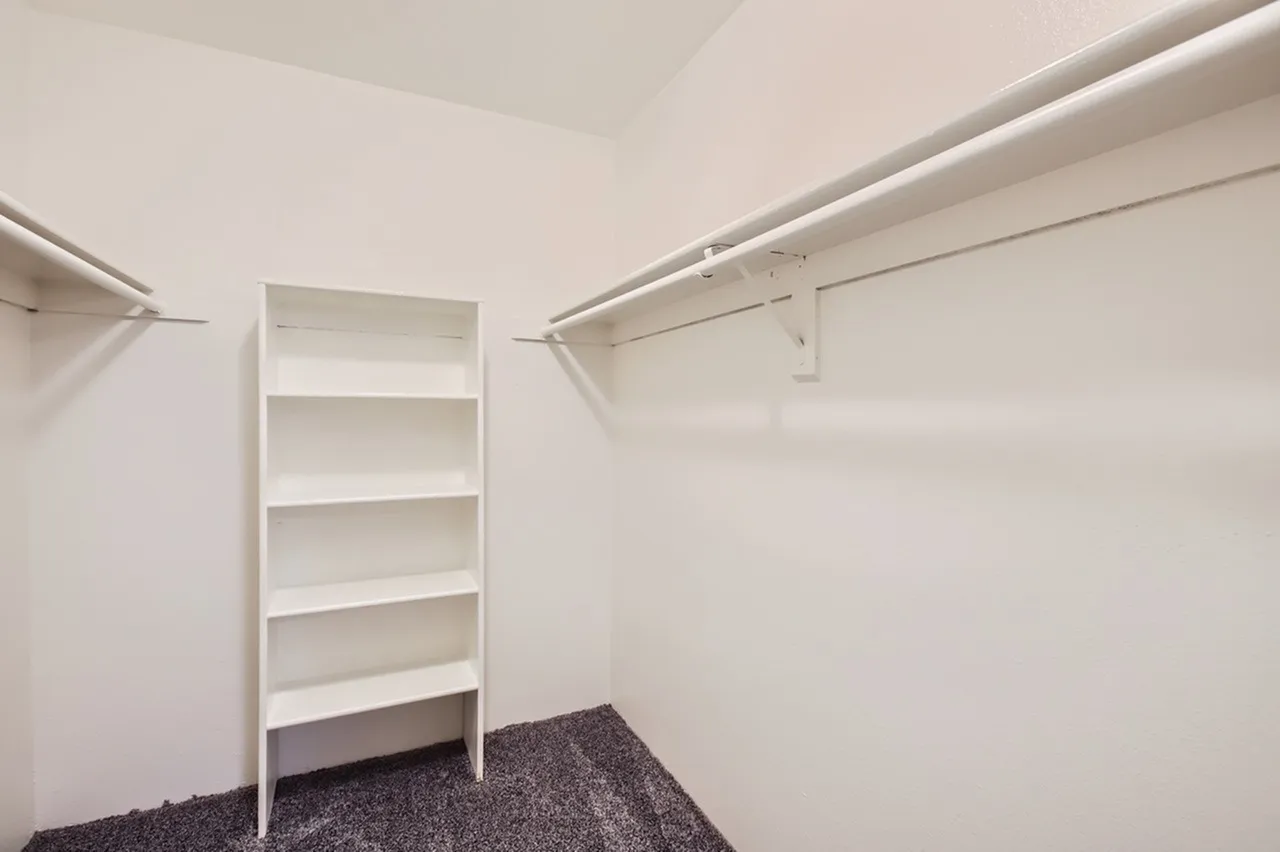 Walk-in closet, white shelving unit, hanging rods, gray carpet flooring