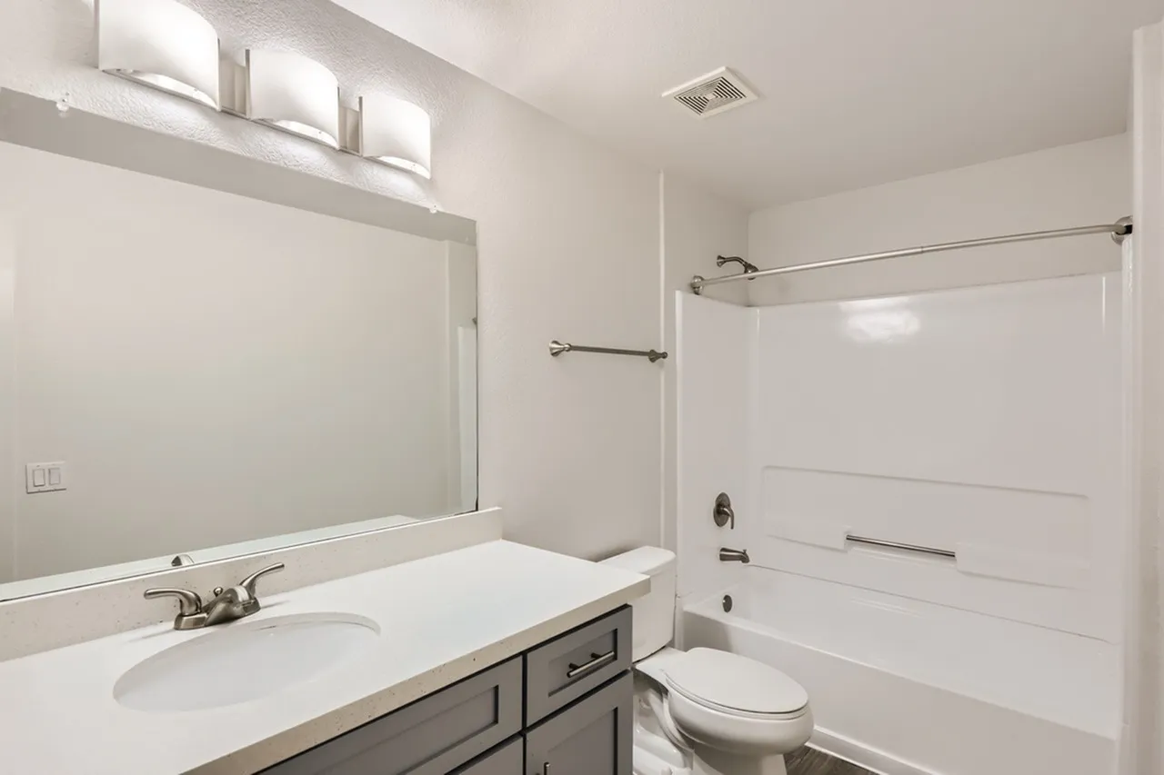 Primary bathroom, white vanity countertop, gray cabinetry, quartz sink, framed mirror, chrome fixtures, white subway tile shower surround, toilet