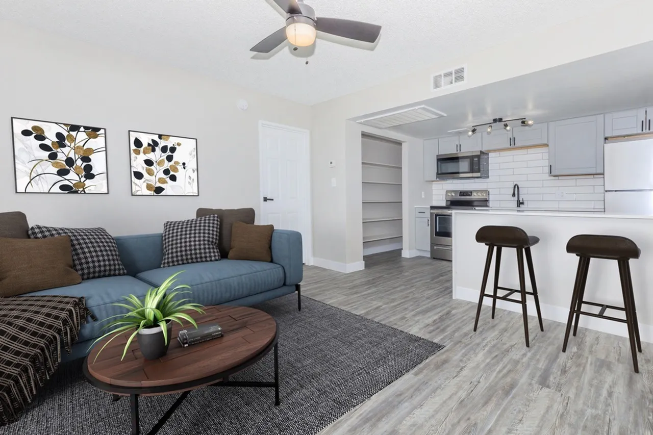 Living room with wood-style plank flooring, blue sofa, ceiling fan, kitchen with white cabinetry and bar seating