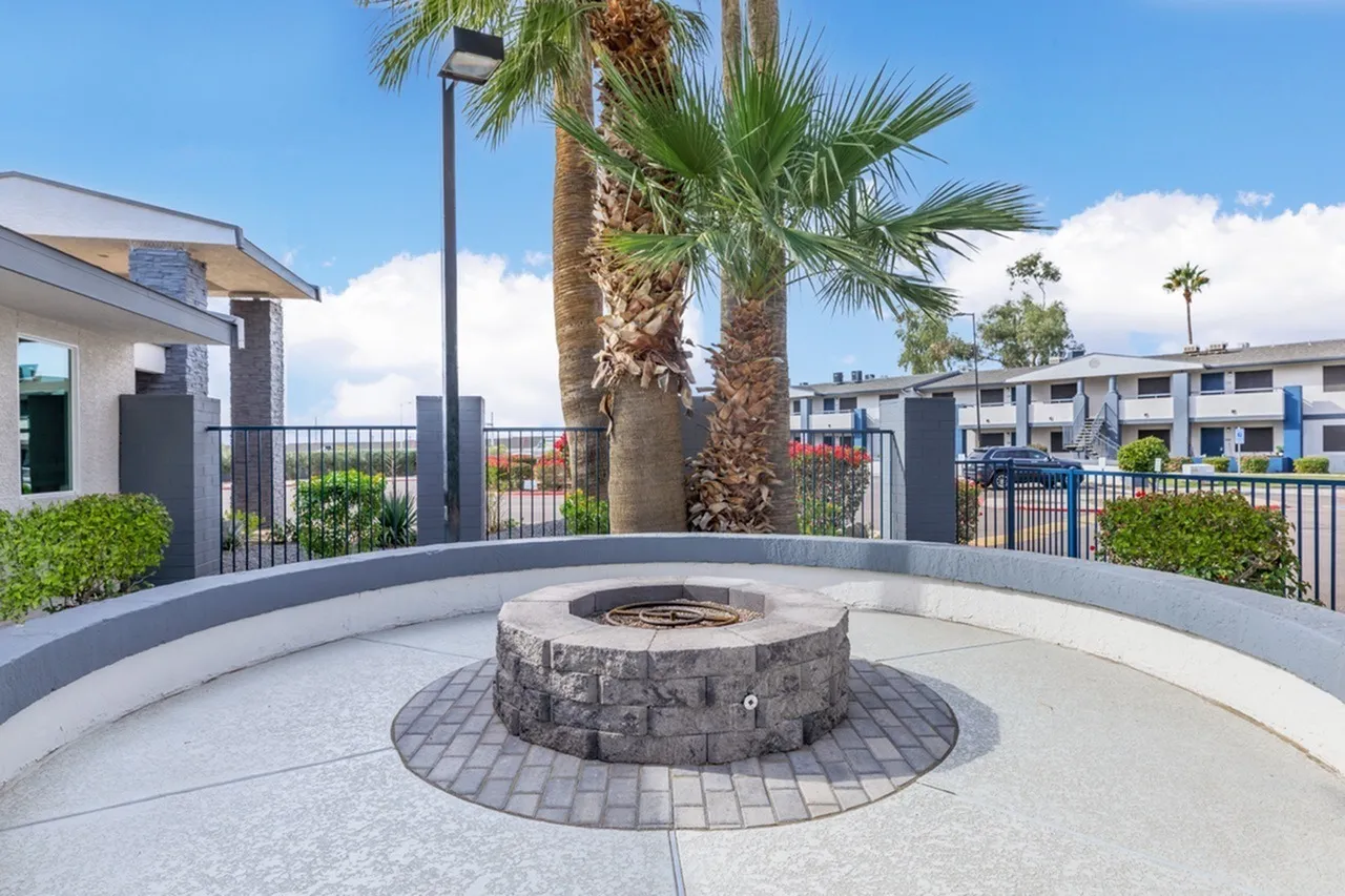 Community fire pit with stone surround, patterned paver base, palm trees, metal railing, apartments in background