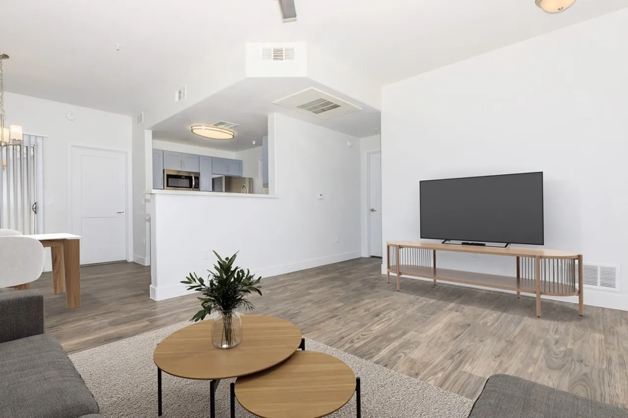 Living room with wood-style plank flooring, round wood coffee tables, flat-screen TV on wooden media console, open kitchen with microwave visible, ceiling fixture, potted plant