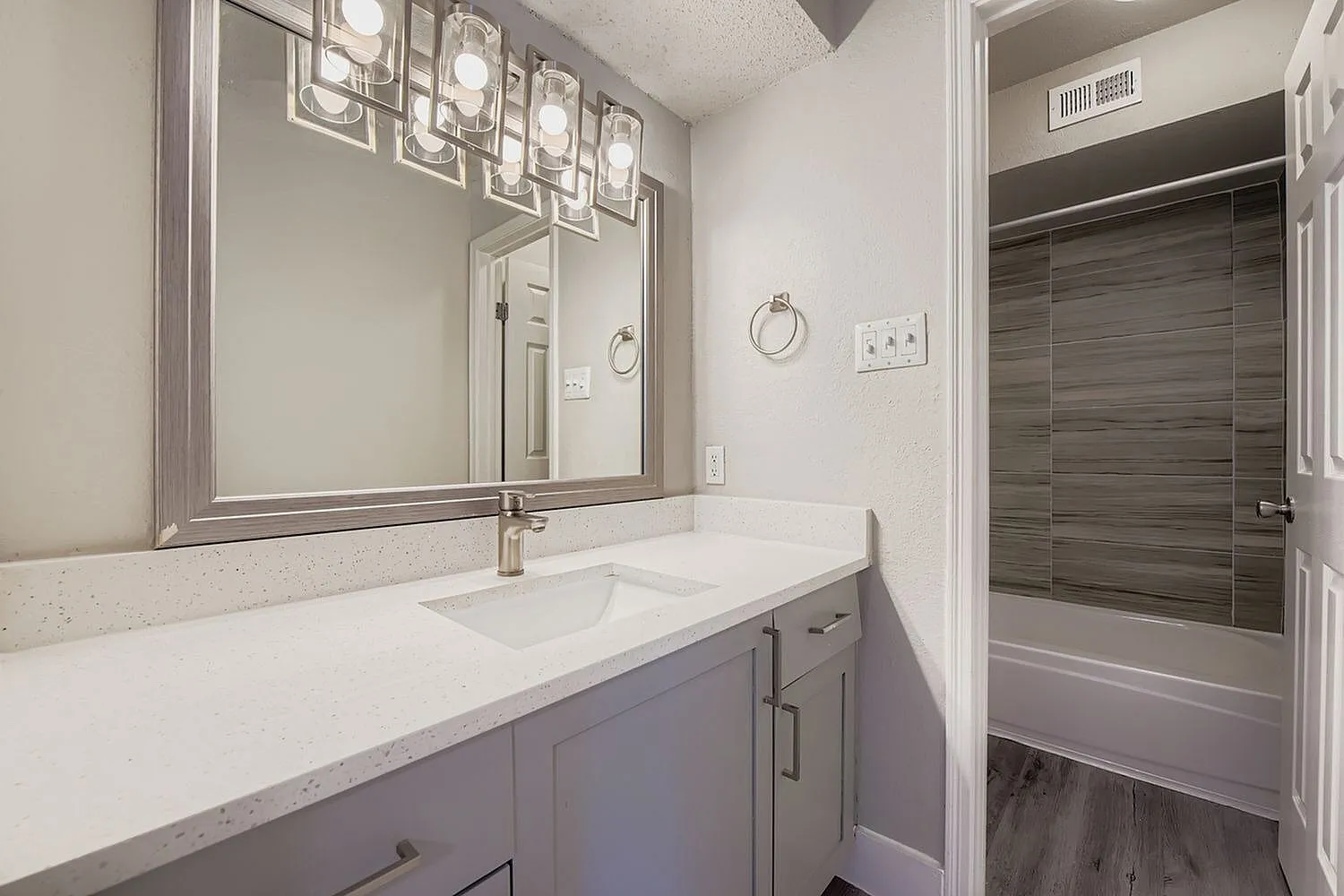 Bathroom vanity, vanity light fixture, framed mirror, quartz-style countertop, wood-look tile tub surround