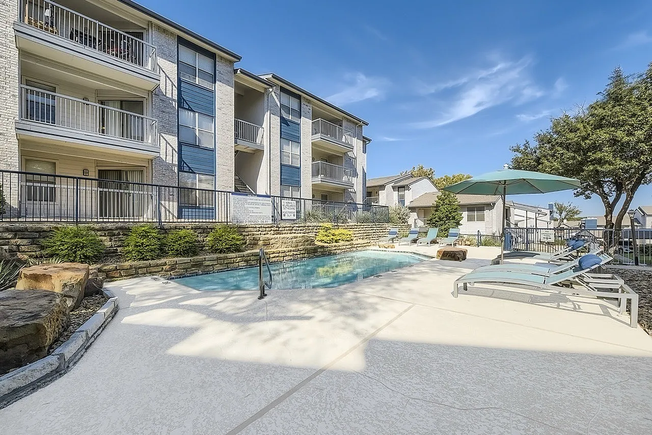 Pool with handrail, stone wall, apartment building, teal patio umbrella
