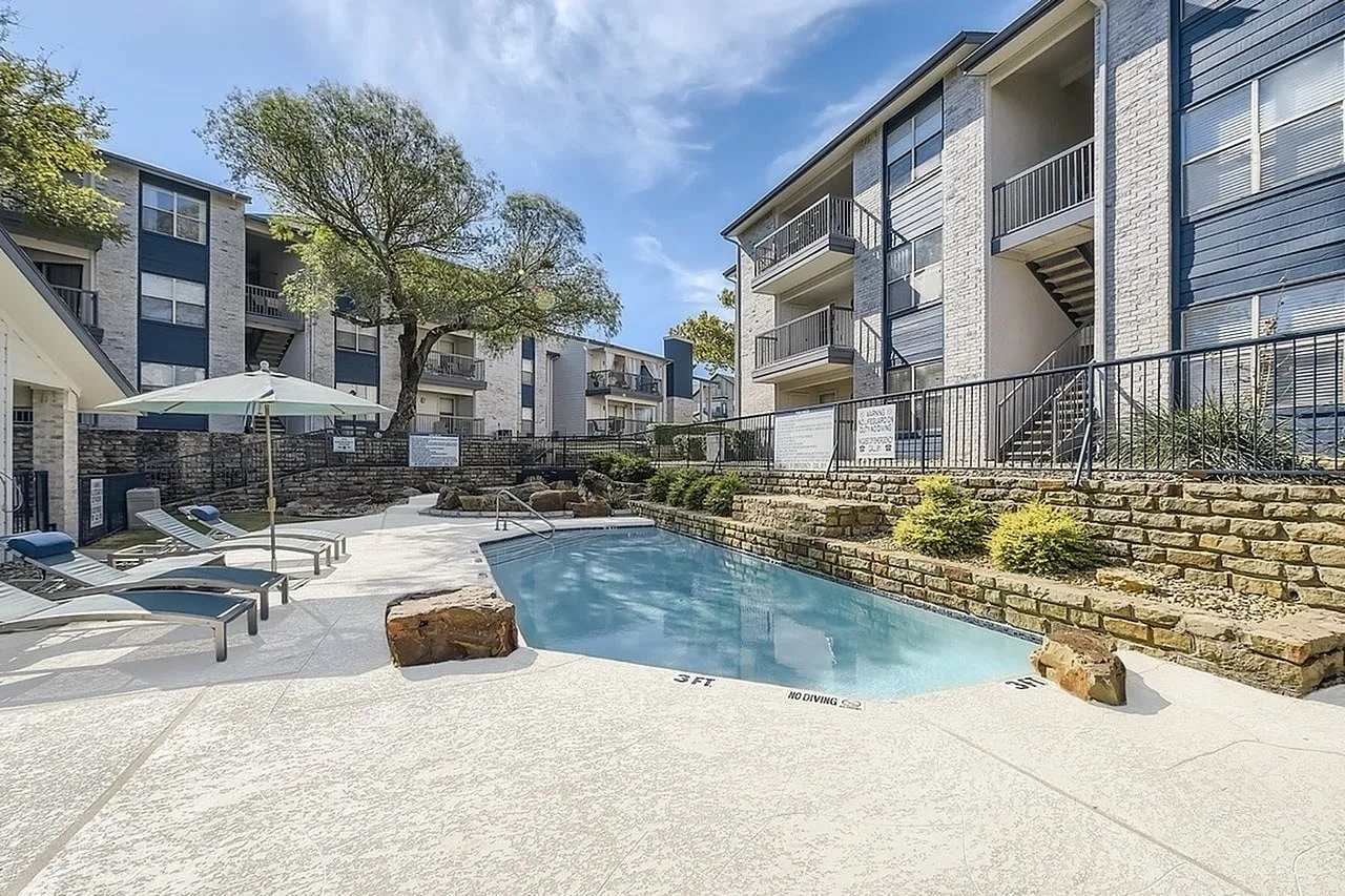 Pool, stone retaining wall, patio umbrella, apartment building with balconies