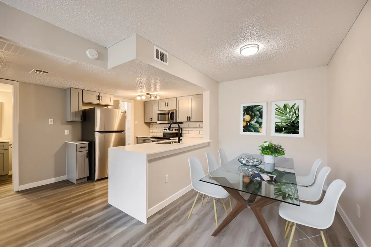 Open concept kitchen and dining area, wood-style plank flooring, stainless steel appliances, white shaker cabinetry, quartz countertops, glass dining table with white chairs