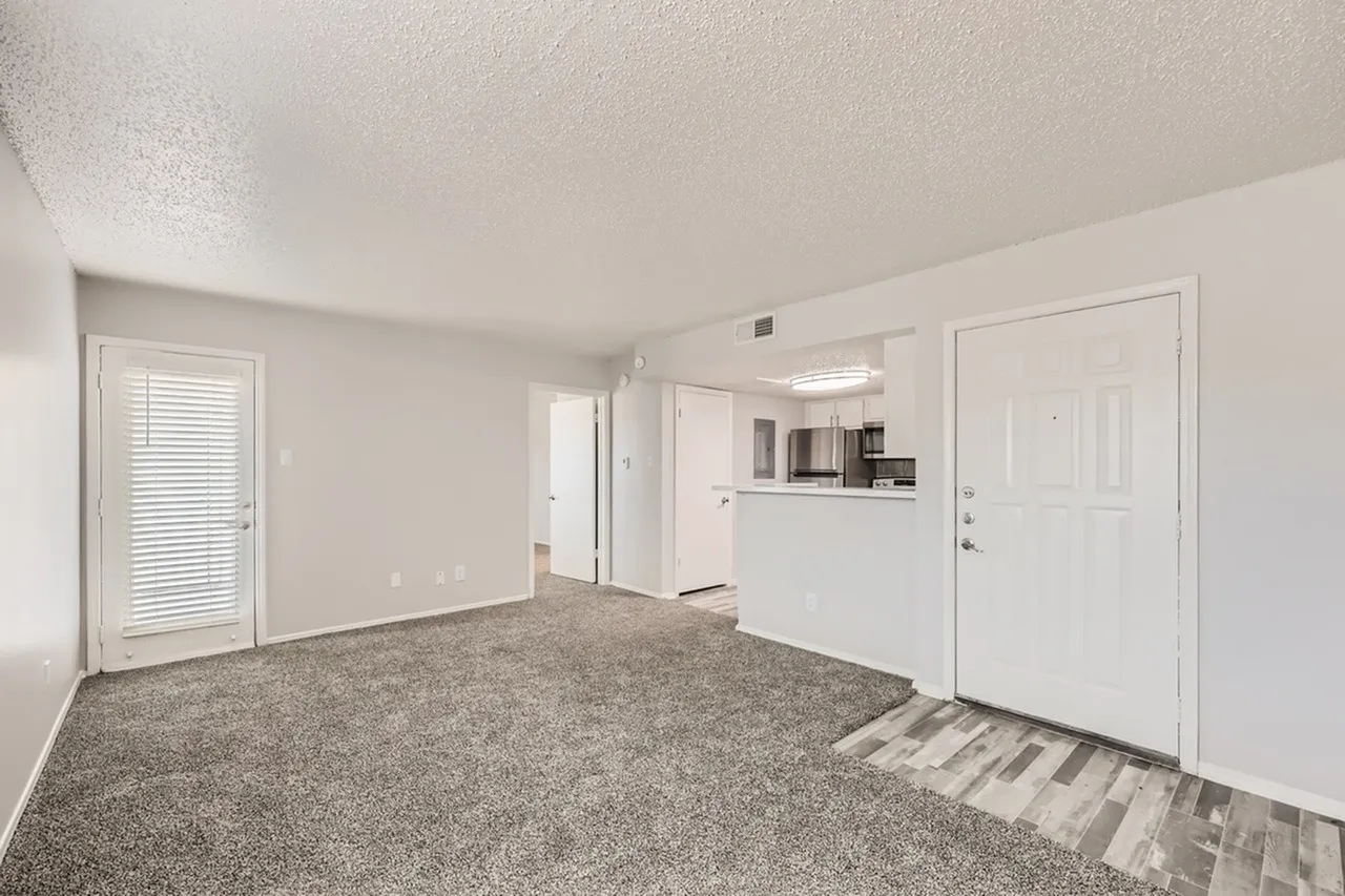 Living room with gray carpet flooring, entry door, open kitchen with white cabinetry visible