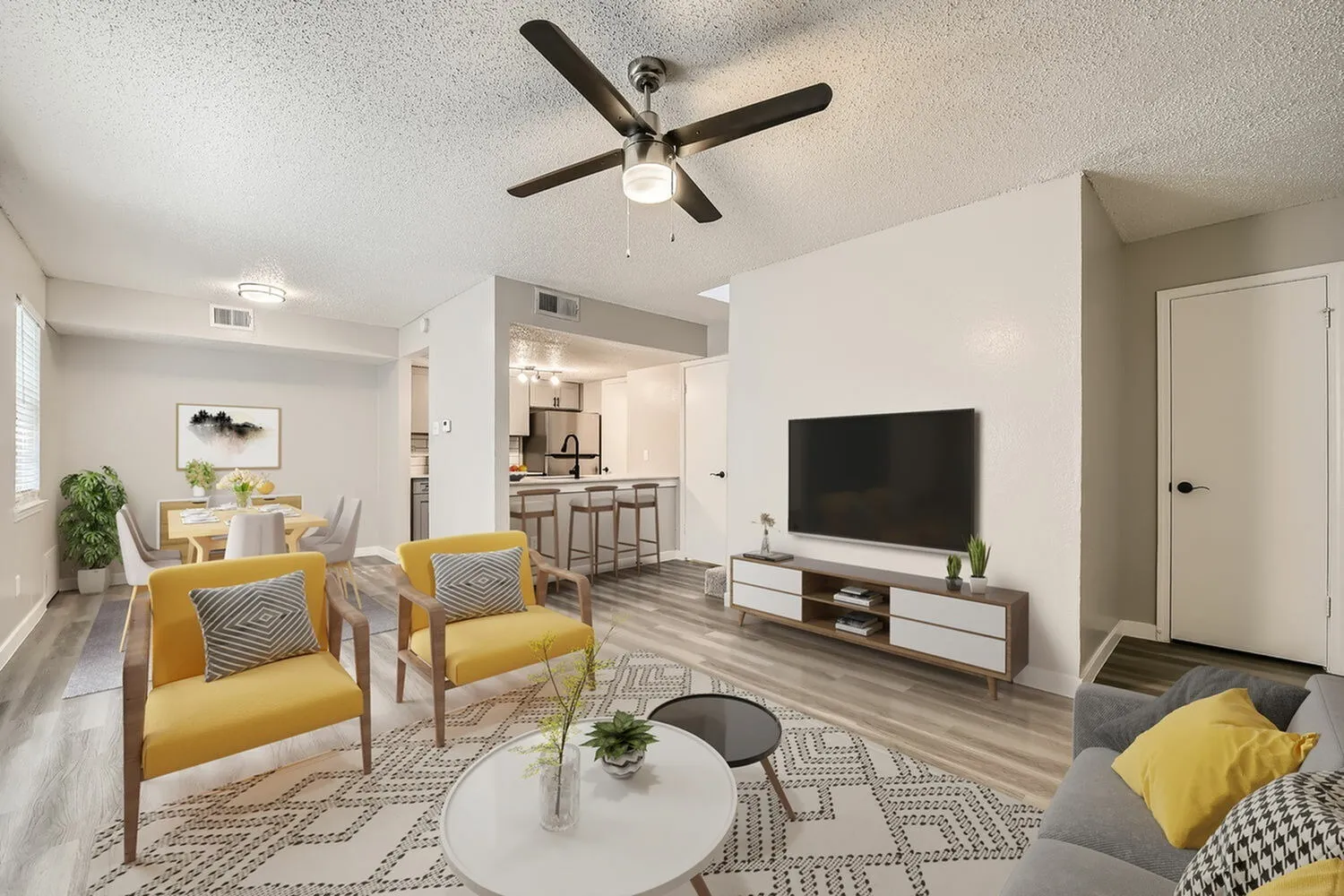 Living room with wood-style plank flooring, ceiling fan, gray upholstered sofa, yellow accent chairs, television, kitchen with bar seating visible in background